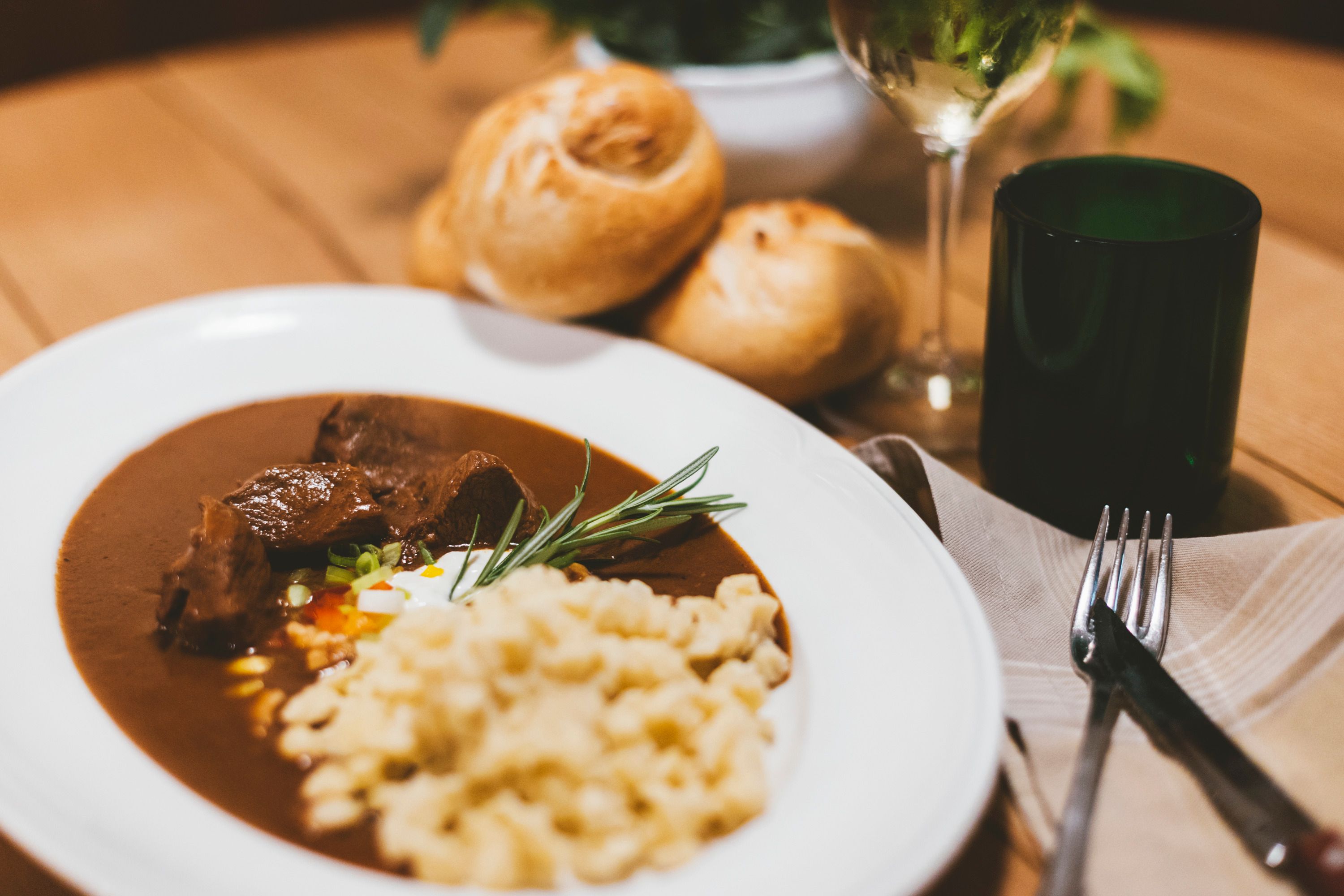 Plate with goulash and spaetzle, bread and wine glass in the background.