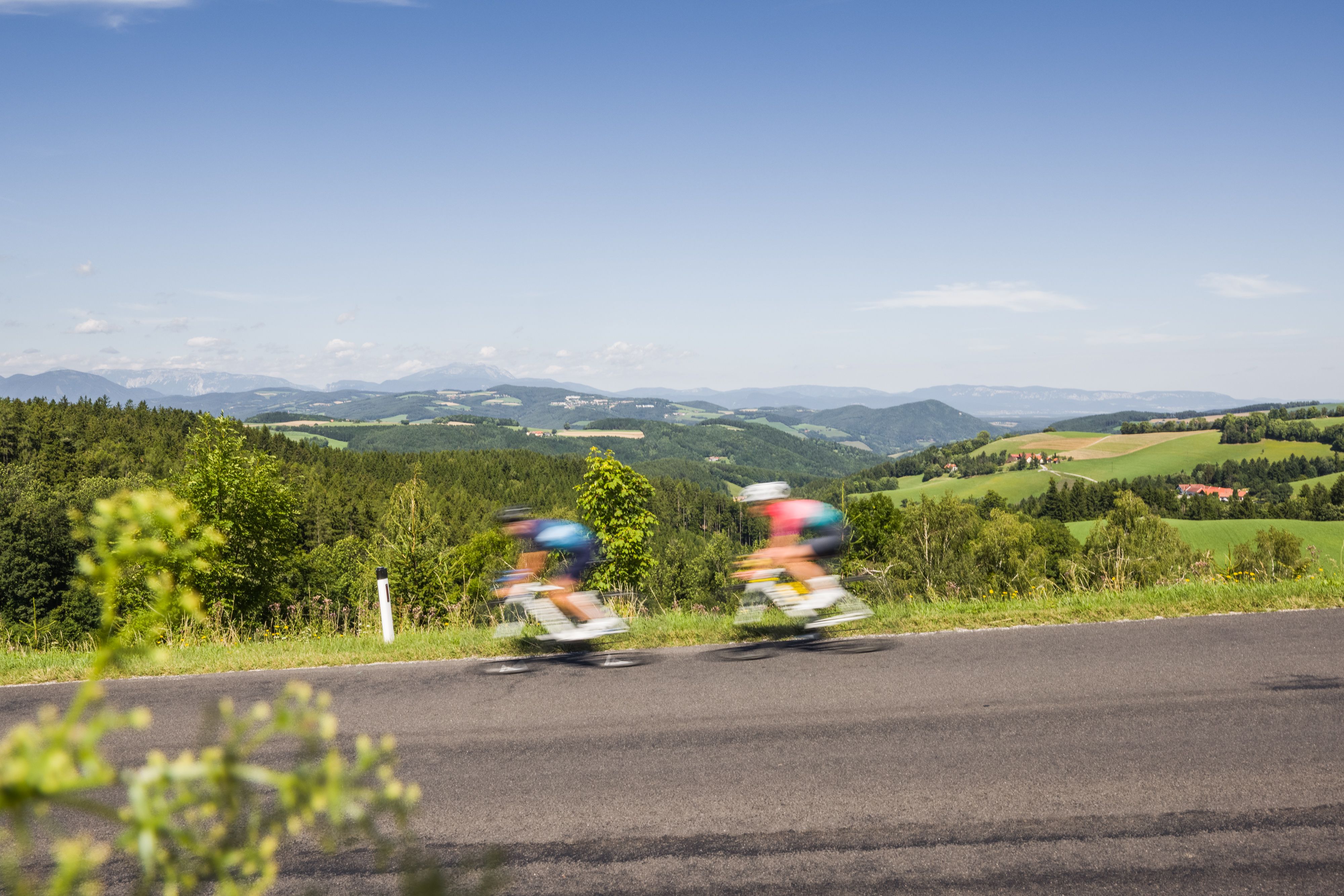 Two people on a road bike riding fast on a road in a hilly landscape.