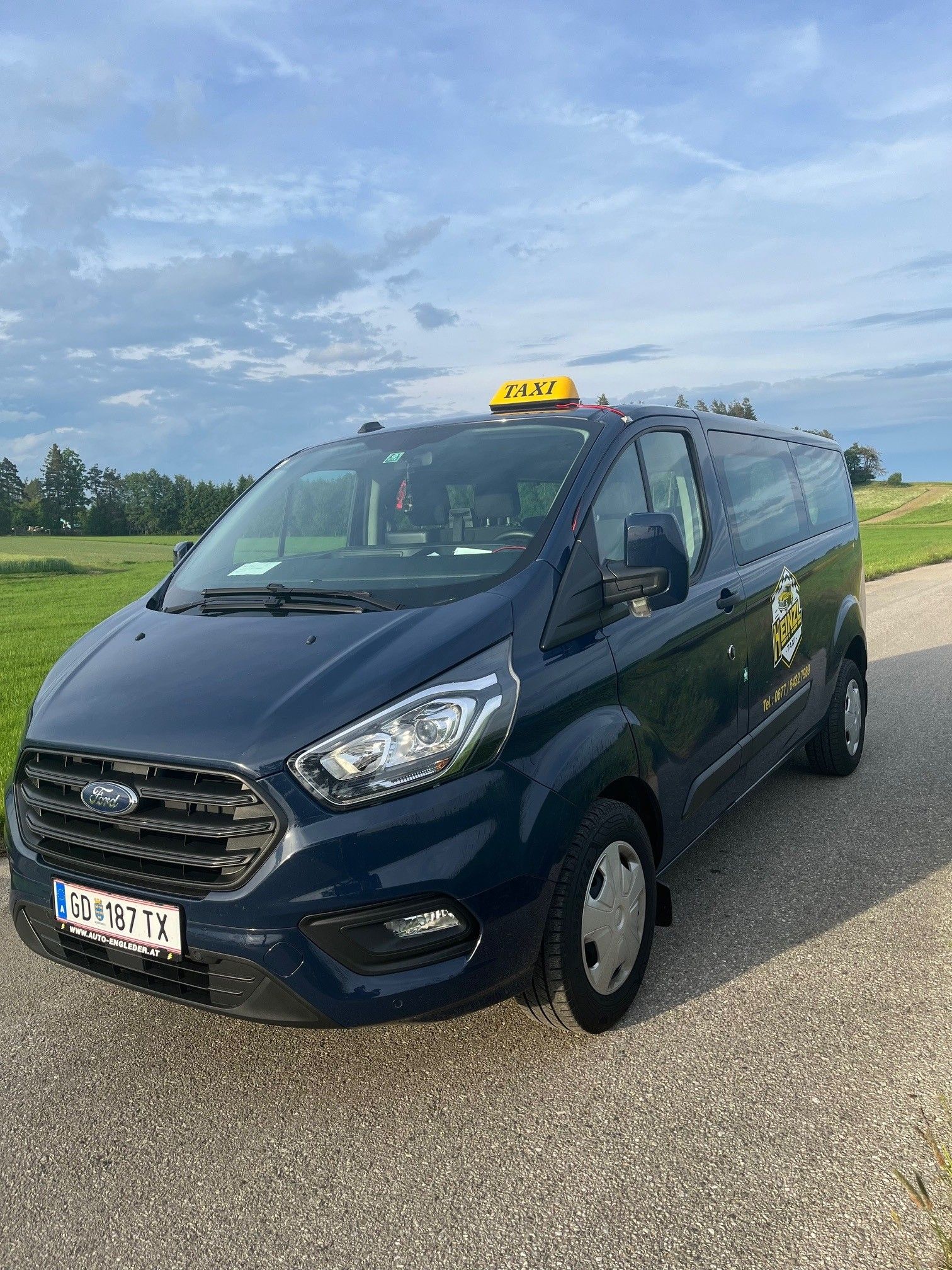 A blue Ford cab is parked on a country road in front of a green meadow and a cloudy sky.