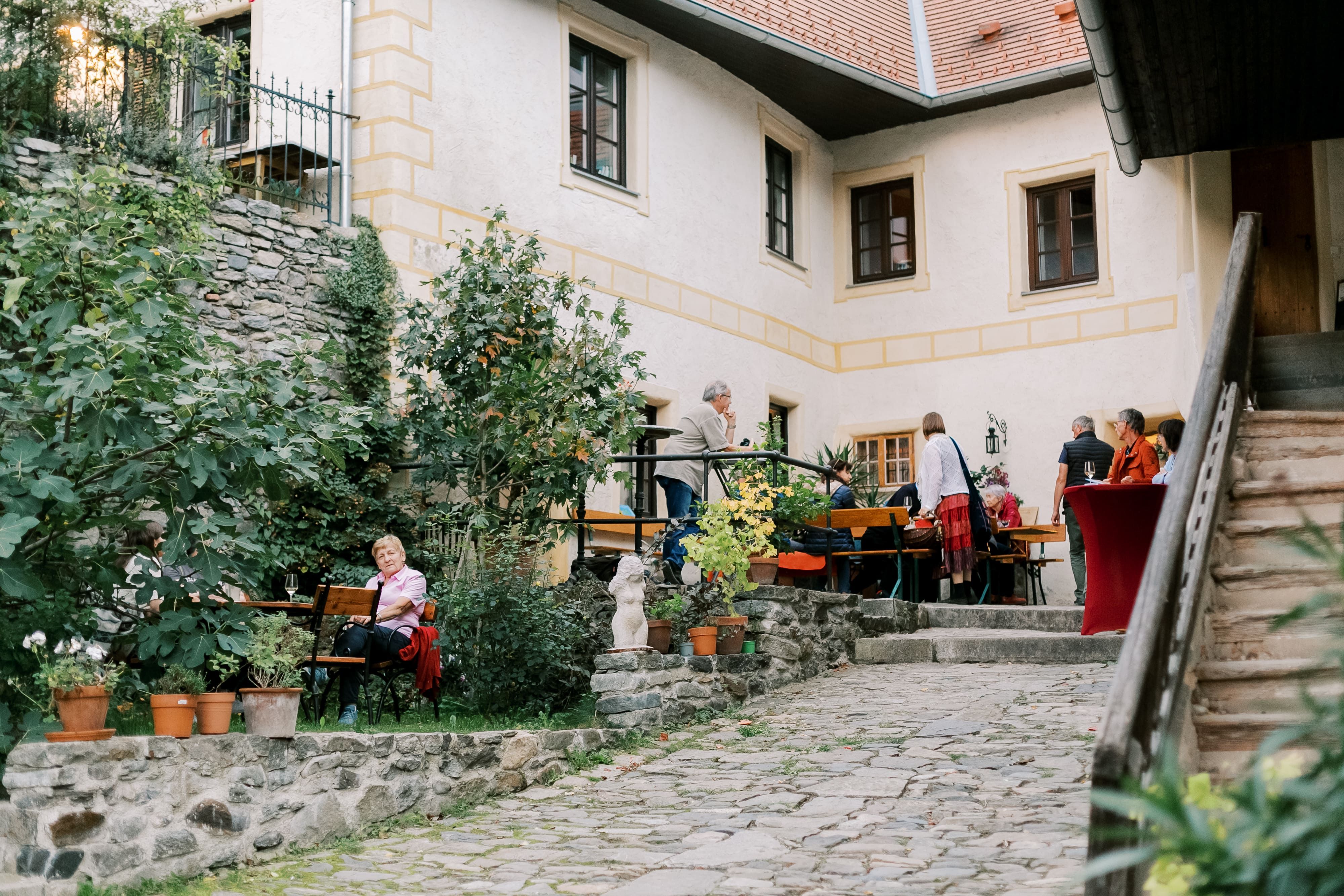An inner courtyard with people sitting and standing at tables, surrounded by plants and a stone building.