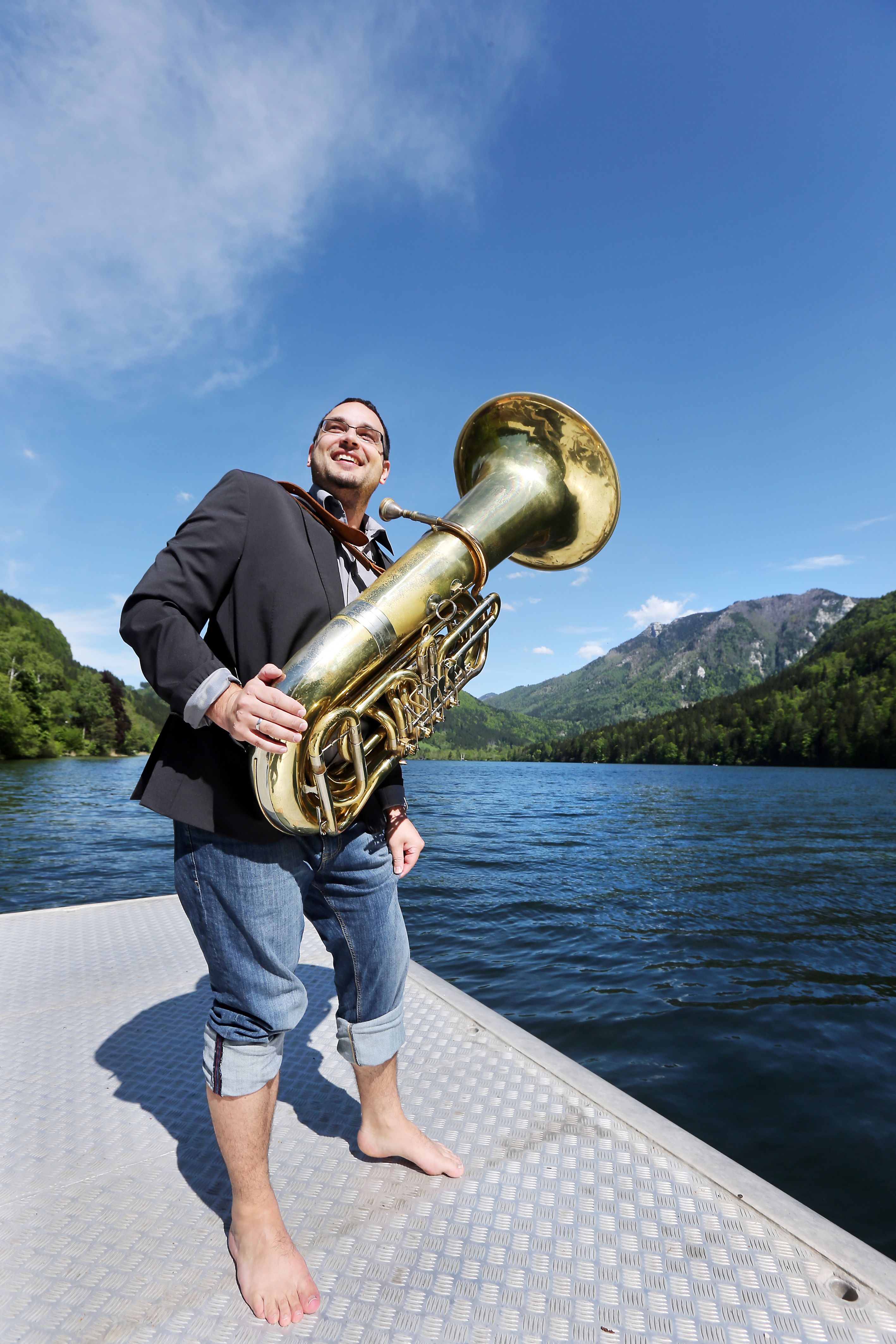 A man with a tuba stands barefoot on a jetty by a lake, surrounded by mountains and blue sky.
