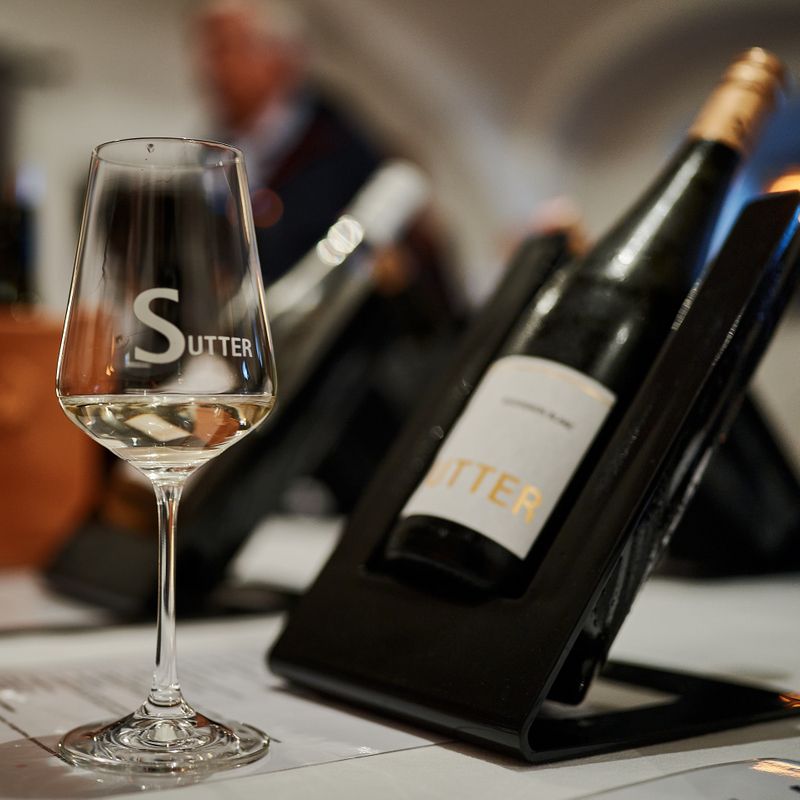 A wine glass labeled 'Sutter' stands in front of a bottle of wine on a table.