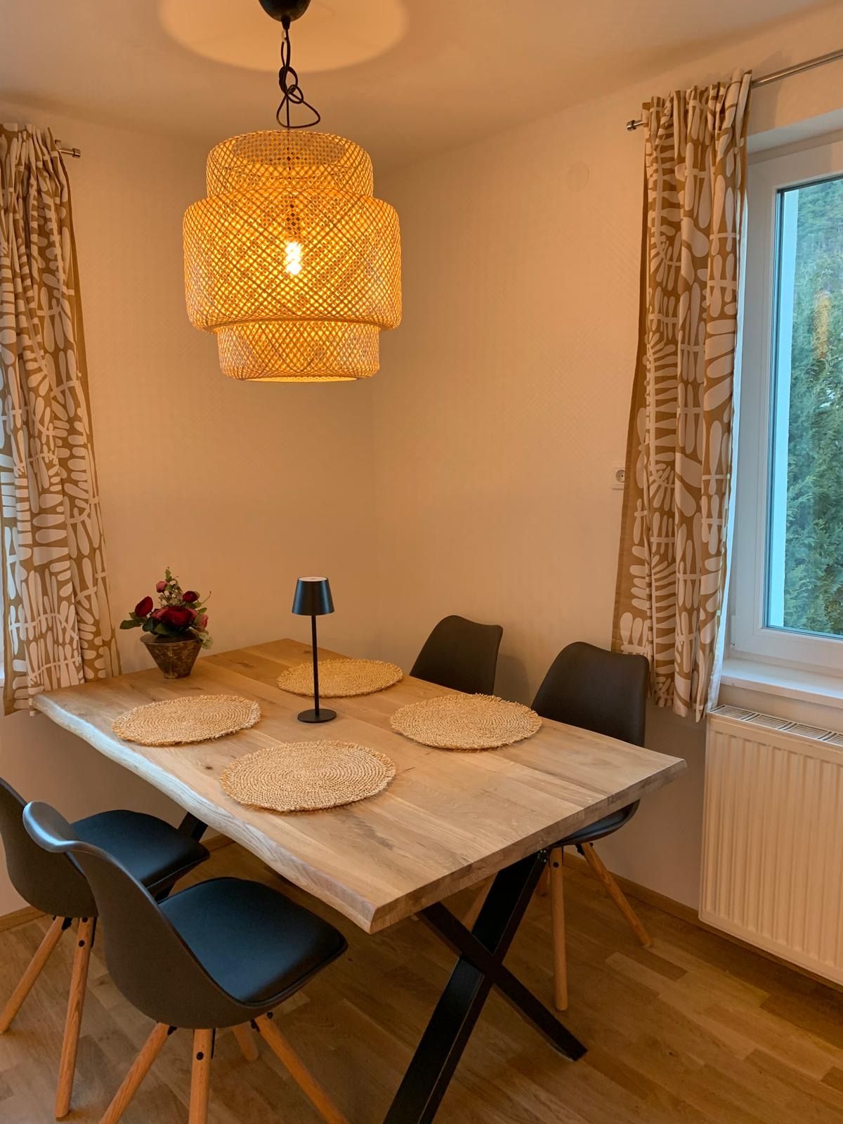 Dining area with wooden table, four black chairs, round lamp and window with curtains.