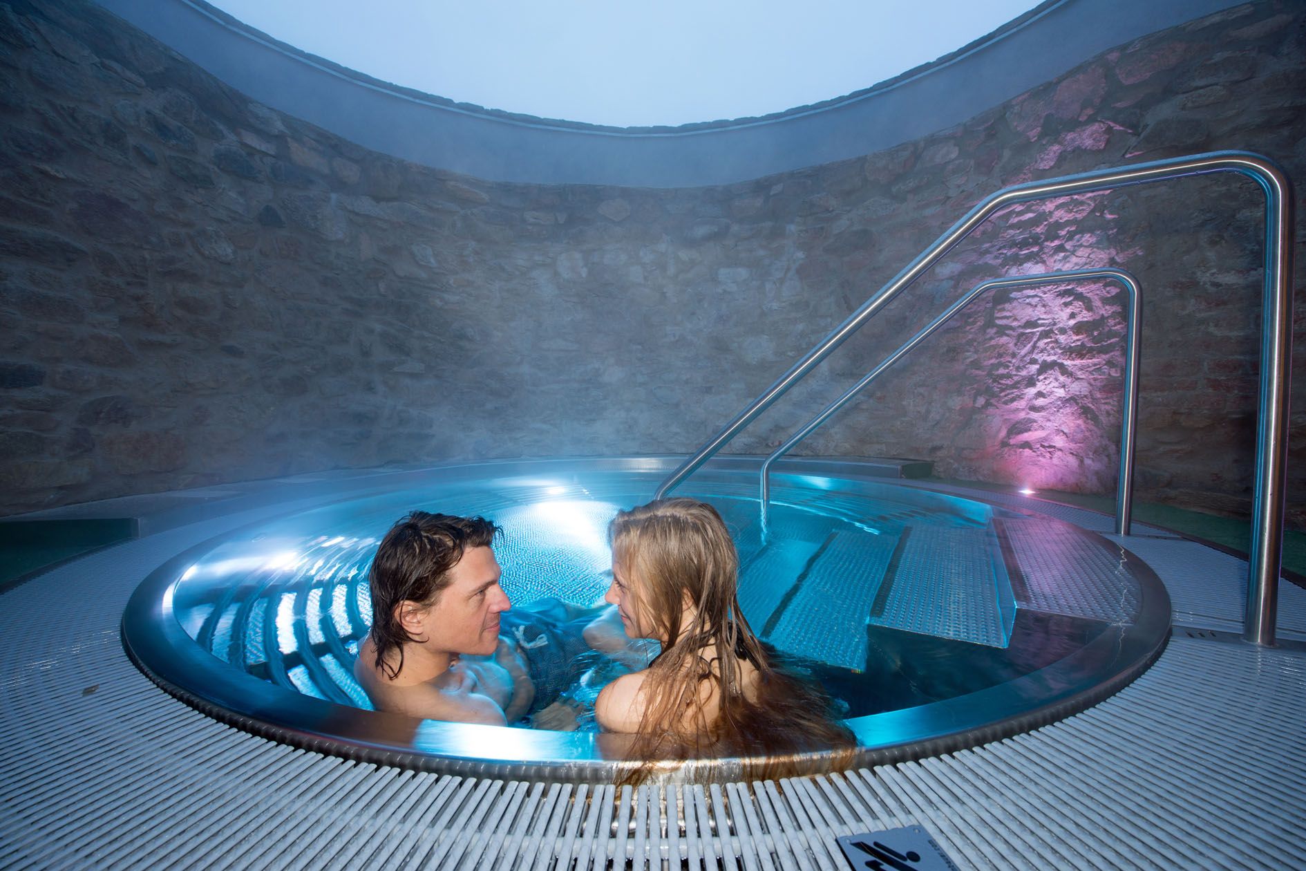 A couple relaxes in a round whirlpool with blue lighting and stone walls.
