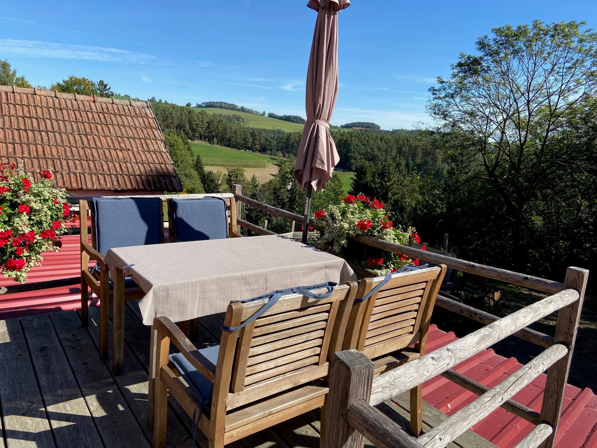Small terrace with wooden furniture, parasol and flower boxes, surrounded by green landscape.