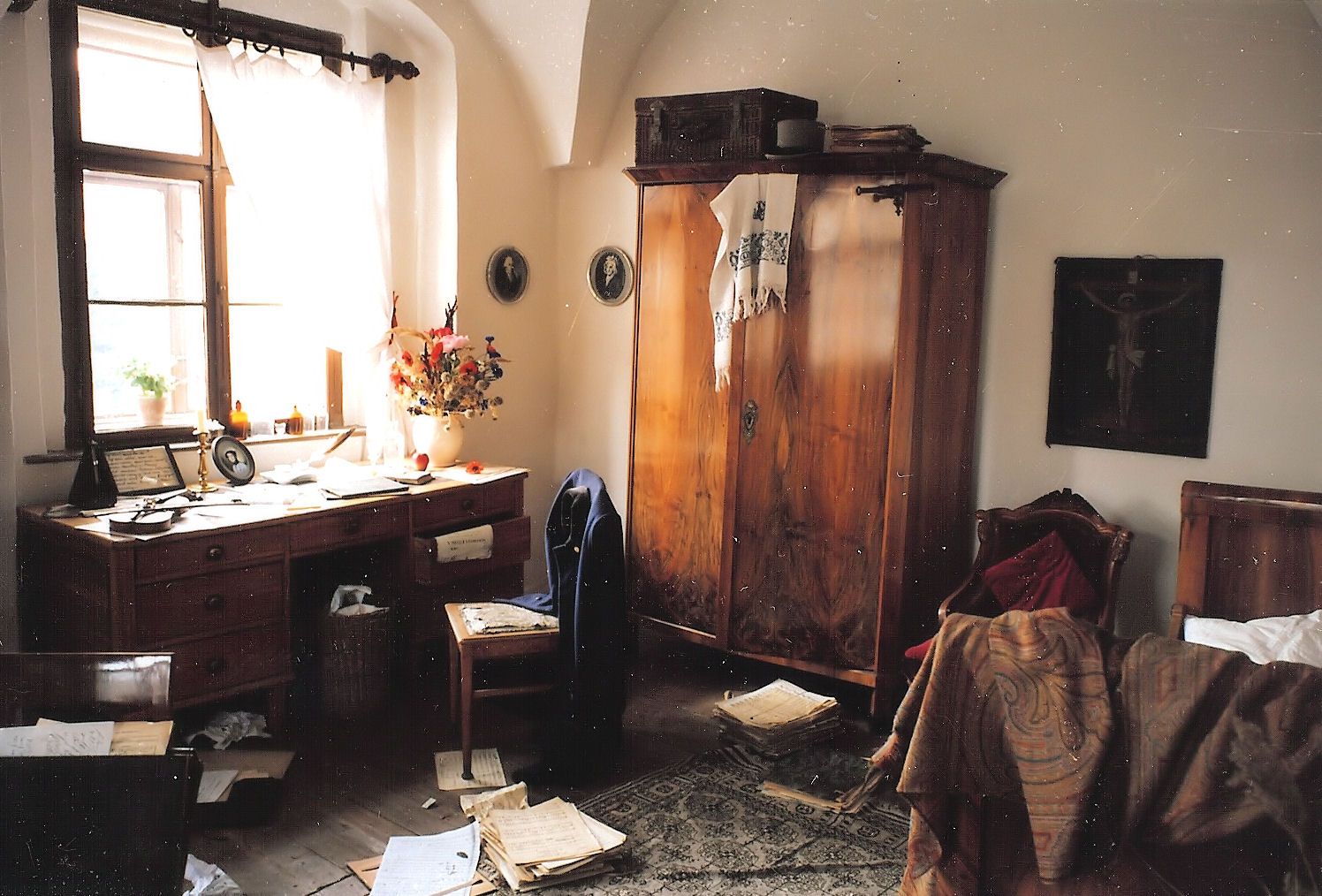 A historic room with a wooden table, sheet music, a wardrobe and a bed. The room looks untidy and old-fashioned.