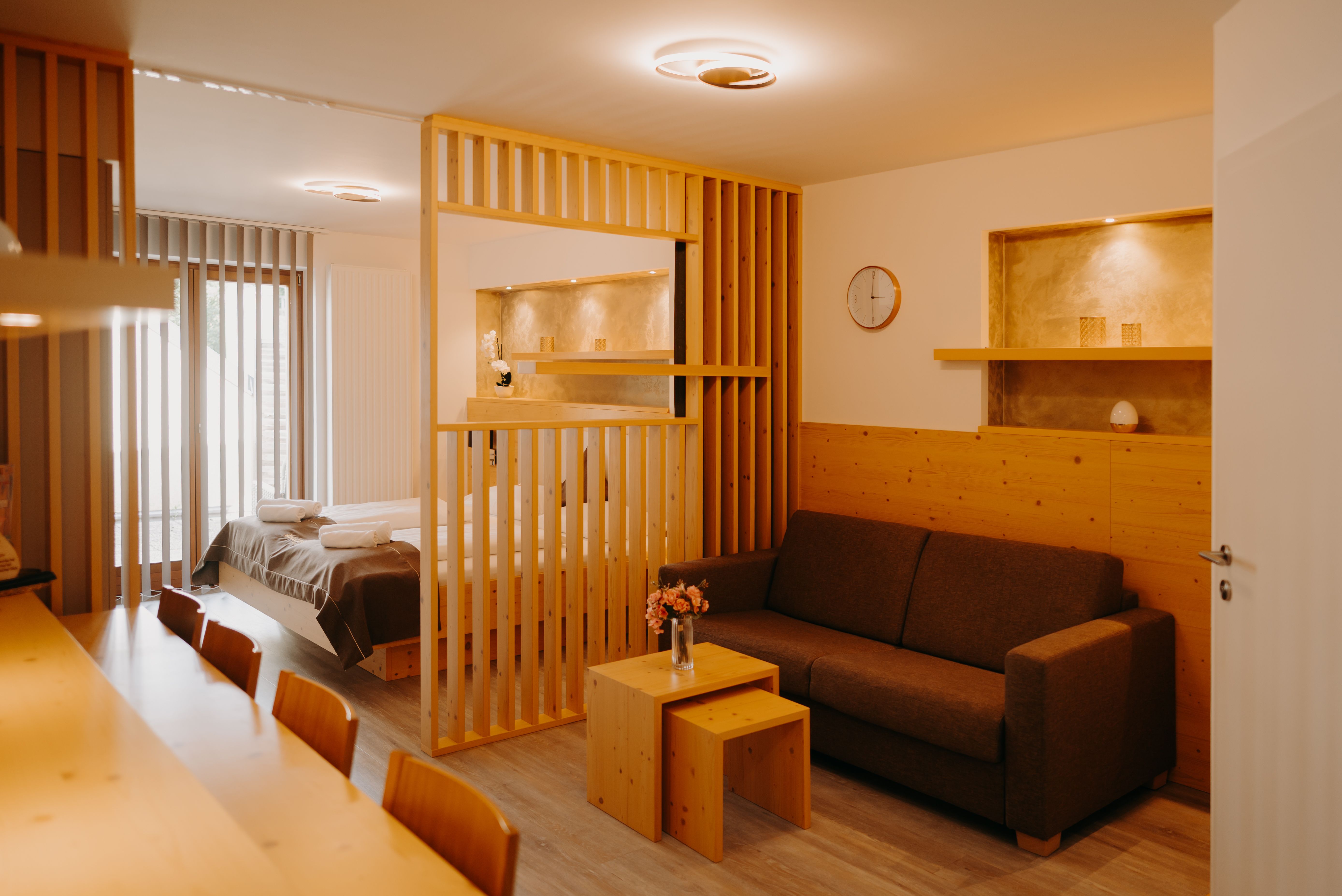 A modern hotel apartment with a double bed, light-colored wood furniture, a pull-out couch, and a wooden room divider. Part of the wall is paneled with wood, and there are niches in the wall painted in an earthy gold.