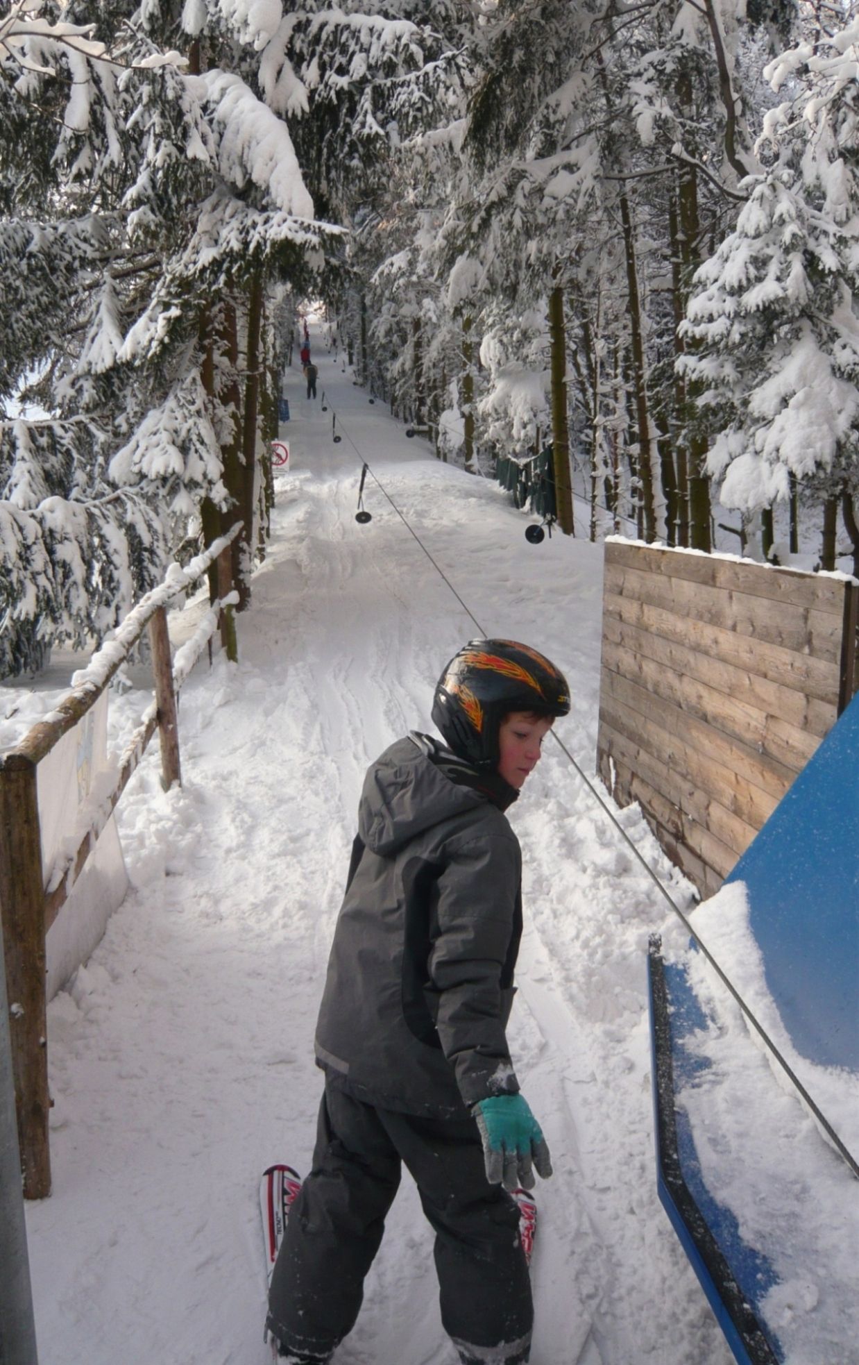 A child wearing a helmet stands on skis at a drag lift in a snowy forest.