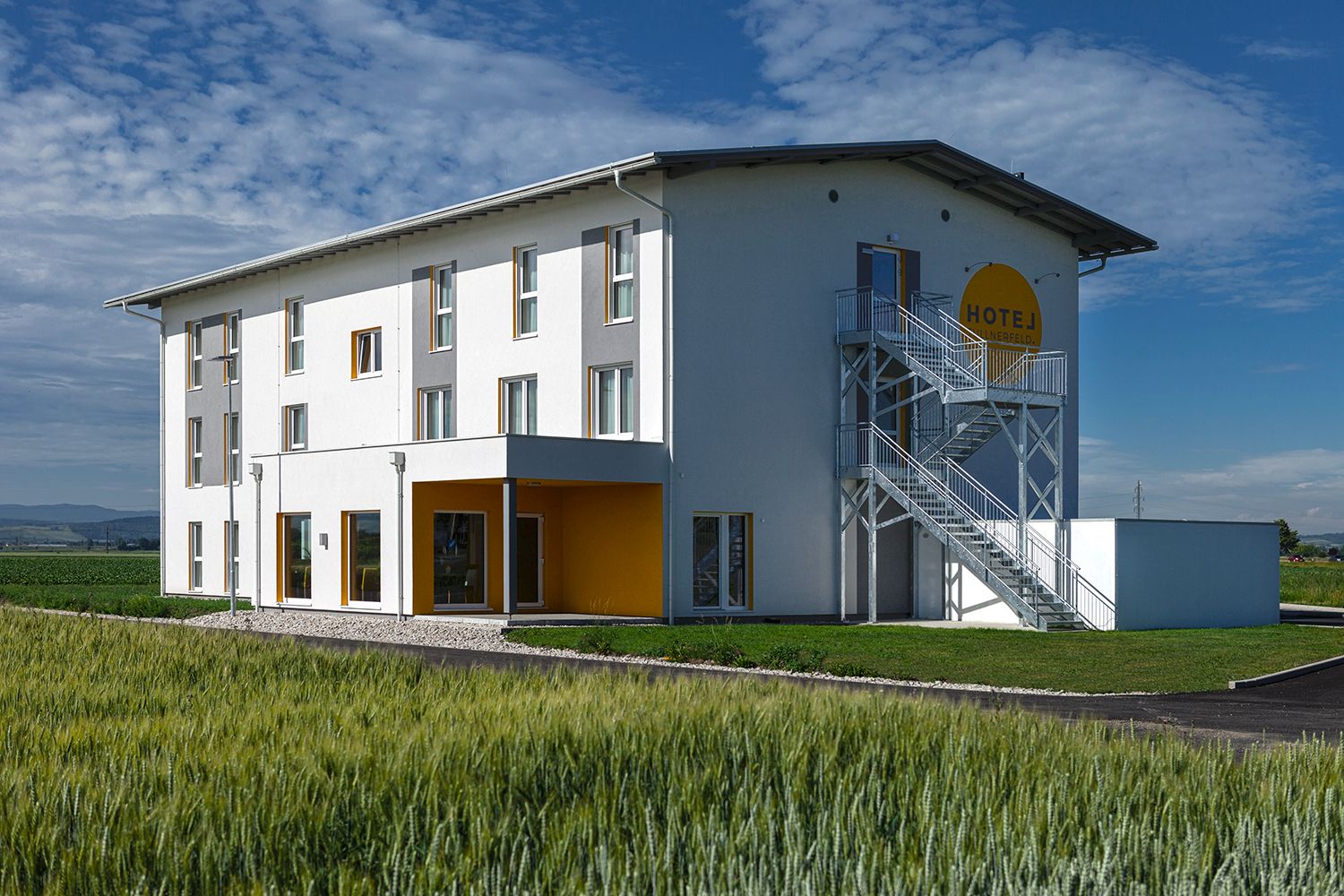 Exterior view of a modern hotel with a white façade and yellow accents, surrounded by fields.