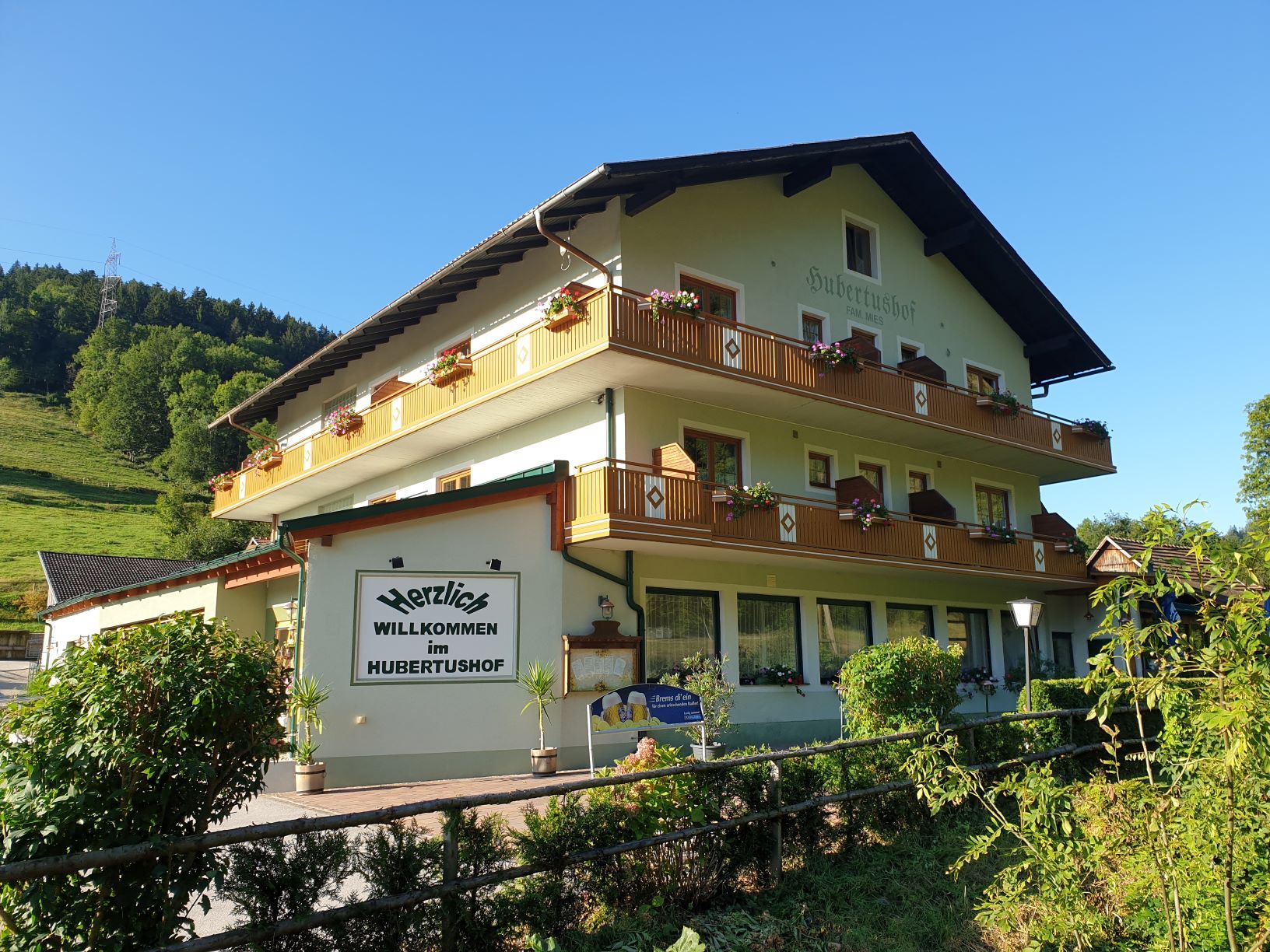 A traditional inn called Hubertushof with flowers on the balconies, surrounded by green countryside and blue skies.