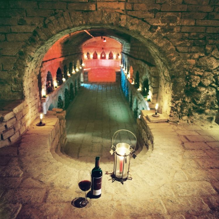An atmospherically lit wine cellar with candles, a bottle of wine and a glass on a stone ledge.