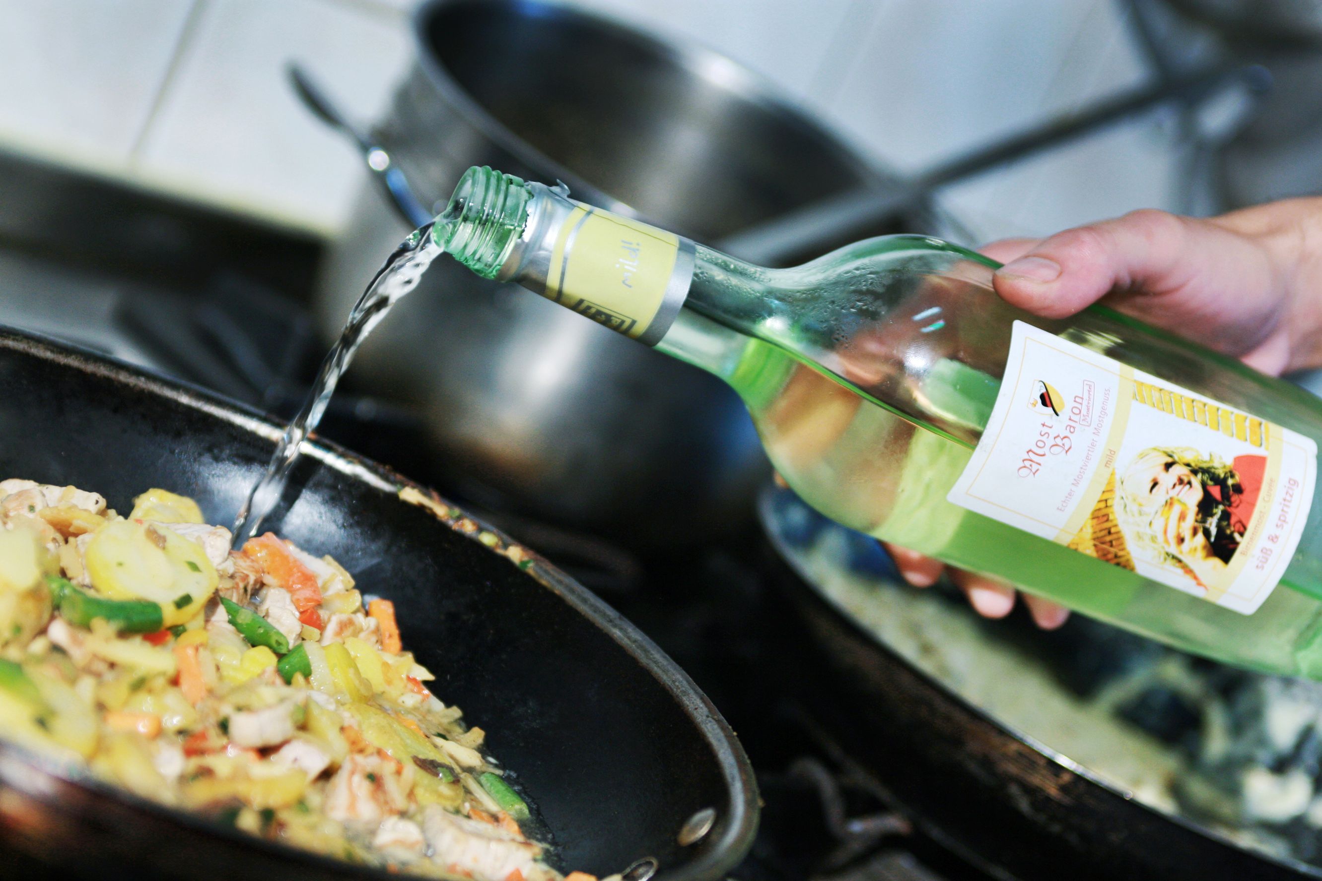 A hand pours Cider from a bottle into a pan of vegetables.