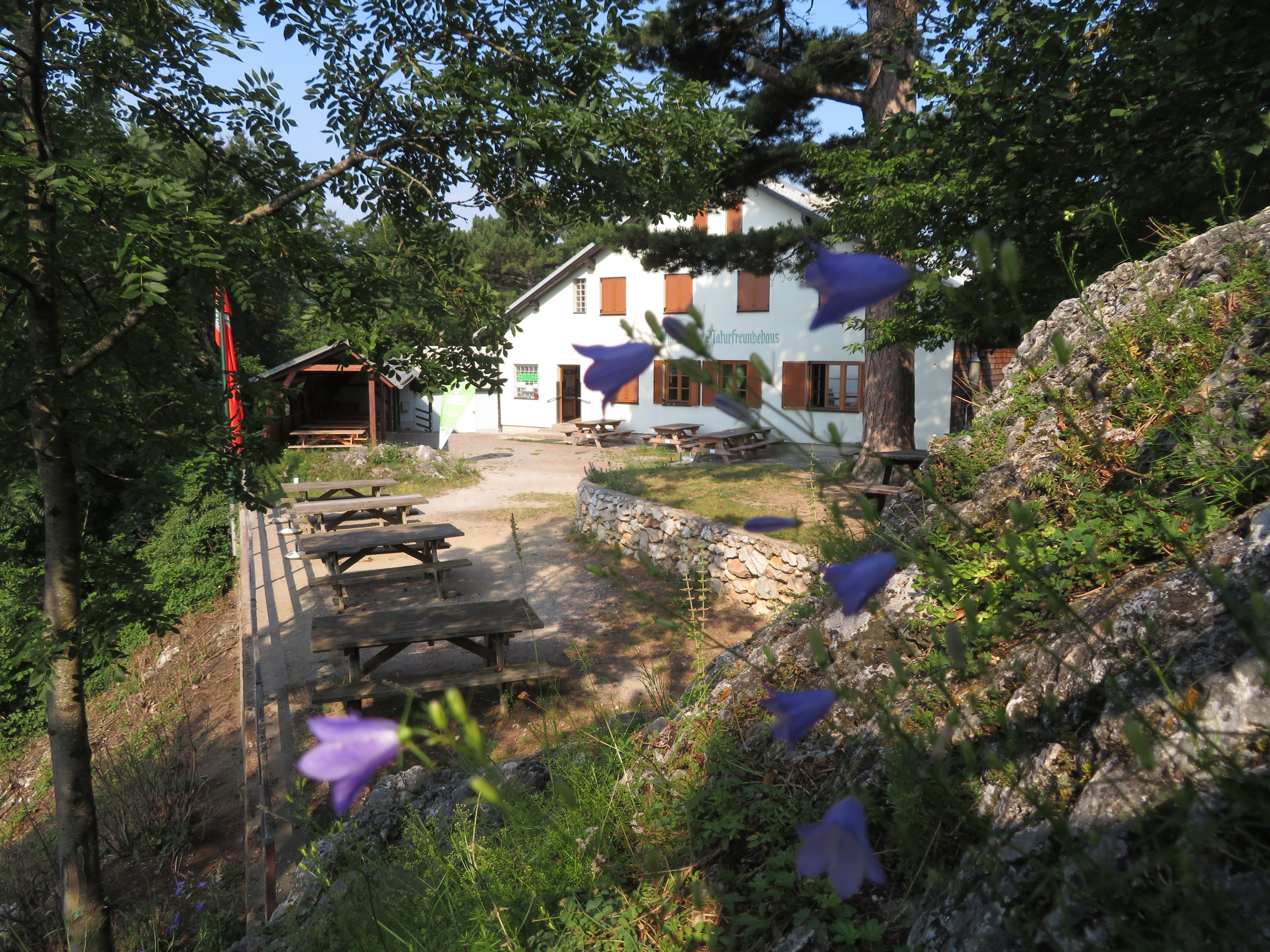 A white building with the inscription 'Naturfreundehaus' surrounded by trees and flowers. Wooden benches and tables can be seen in the foreground.