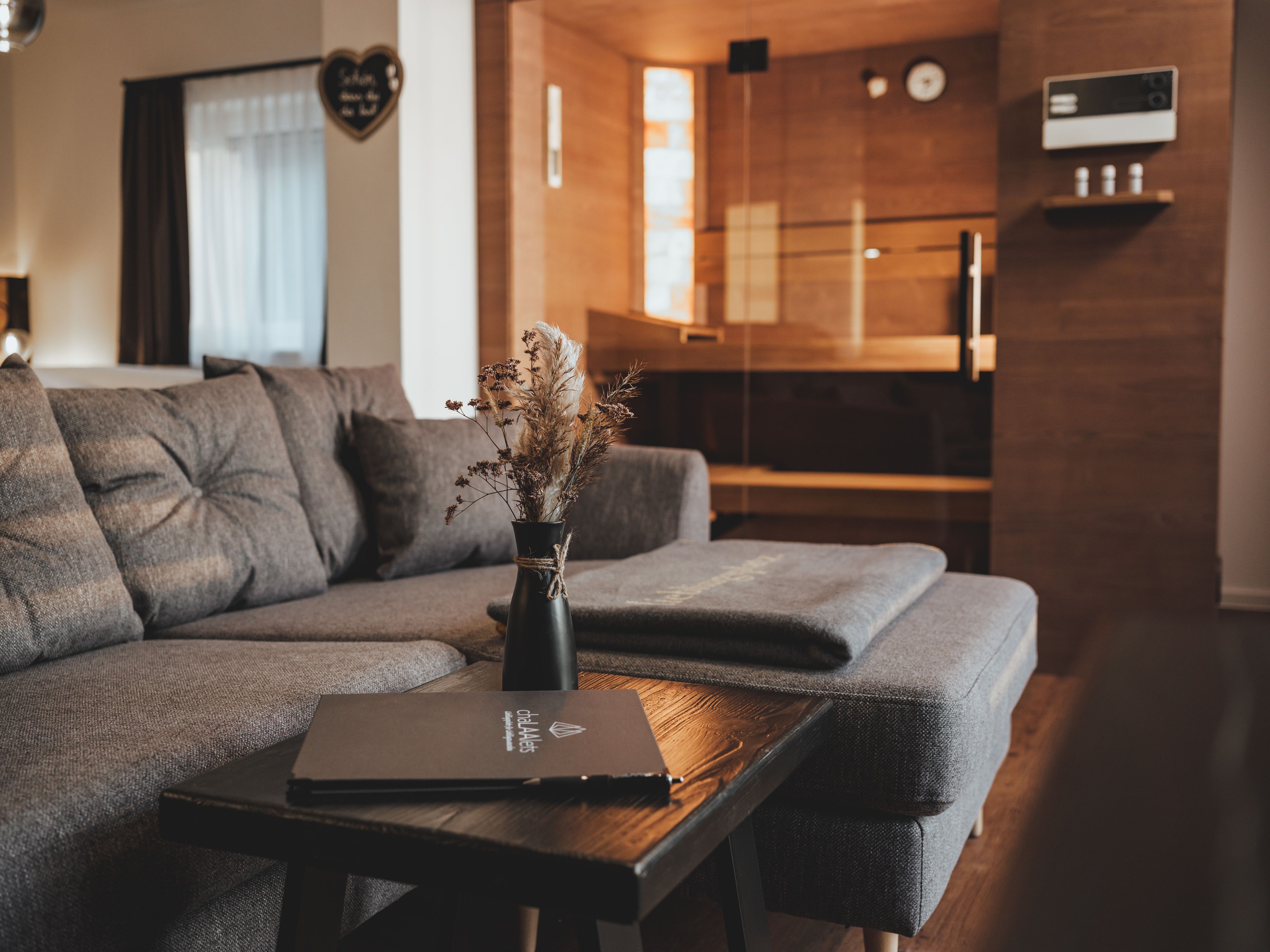 Cozy living room with grey sofa, wooden table and sauna in the background.
