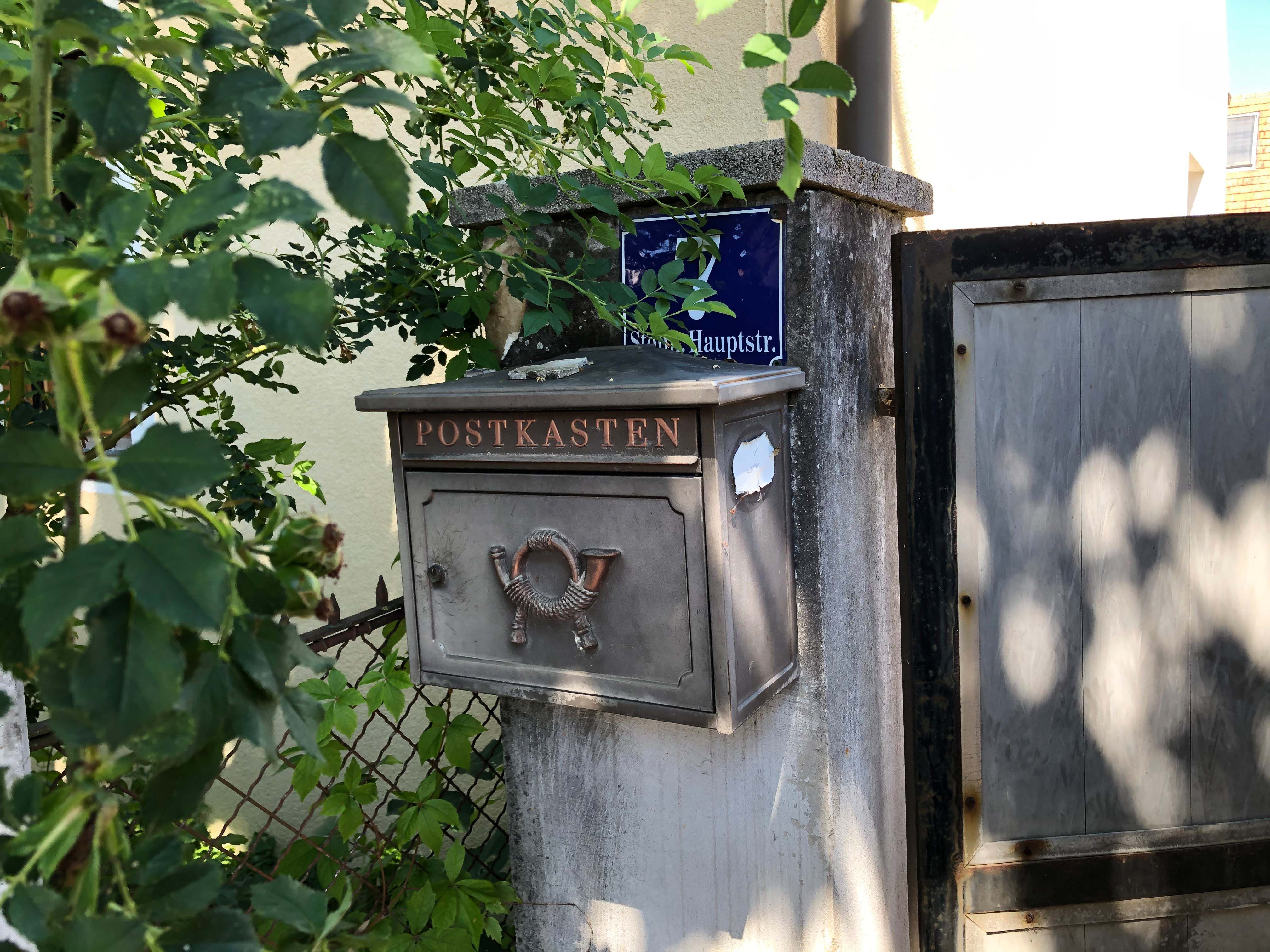 An old letterbox with the inscription 'Postbox' on a wall next to a gate, surrounded by plants.
