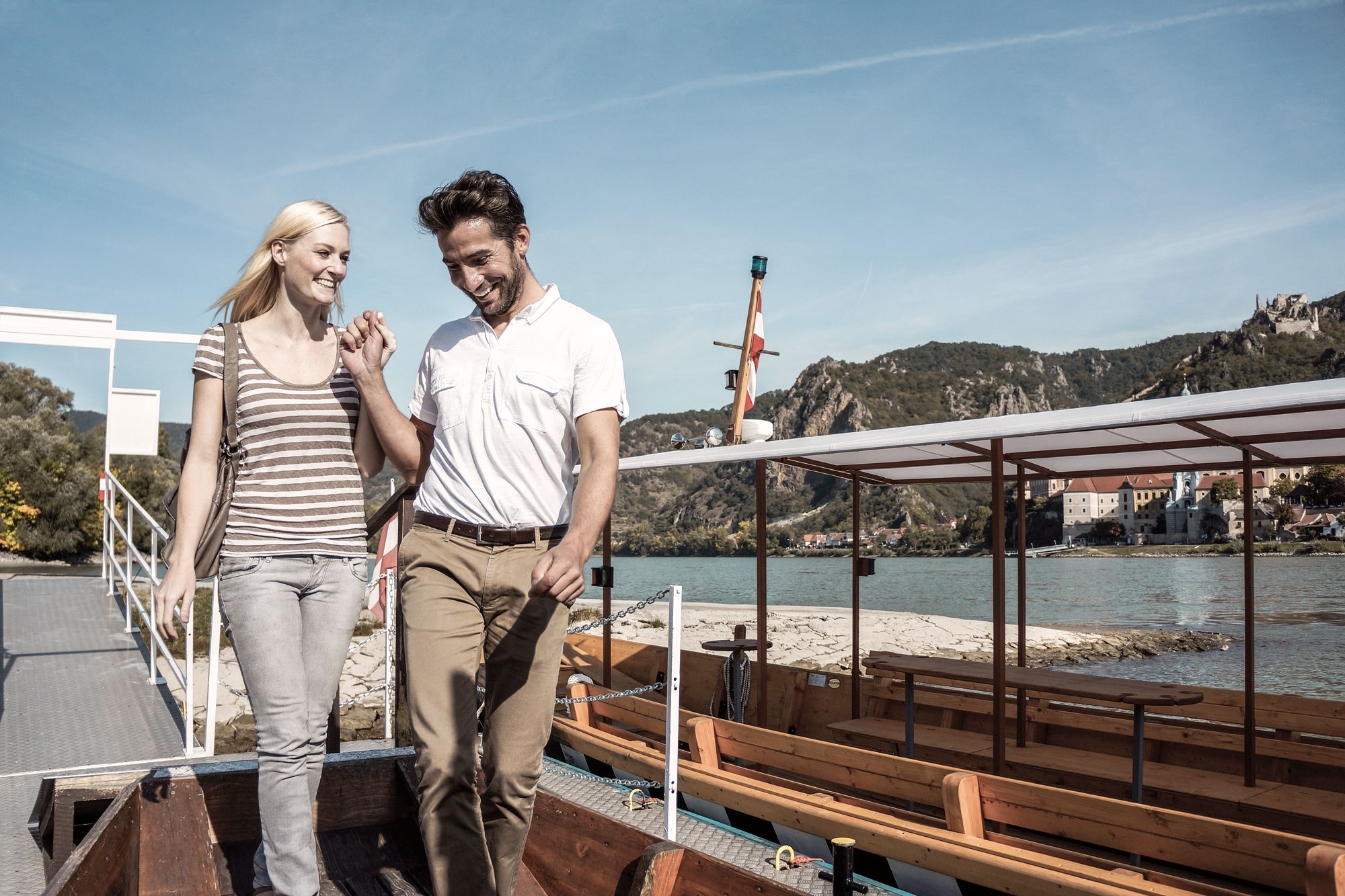 A smiling couple gets out of a boat on a riverbank, mountains and buildings can be seen in the background.