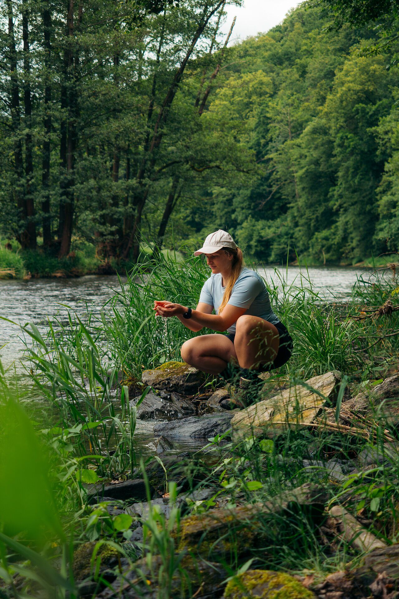 A woman kneels on the banks of the Thaya in the Thayatal National Park, surrounded by dense vegetation and lush greenery.