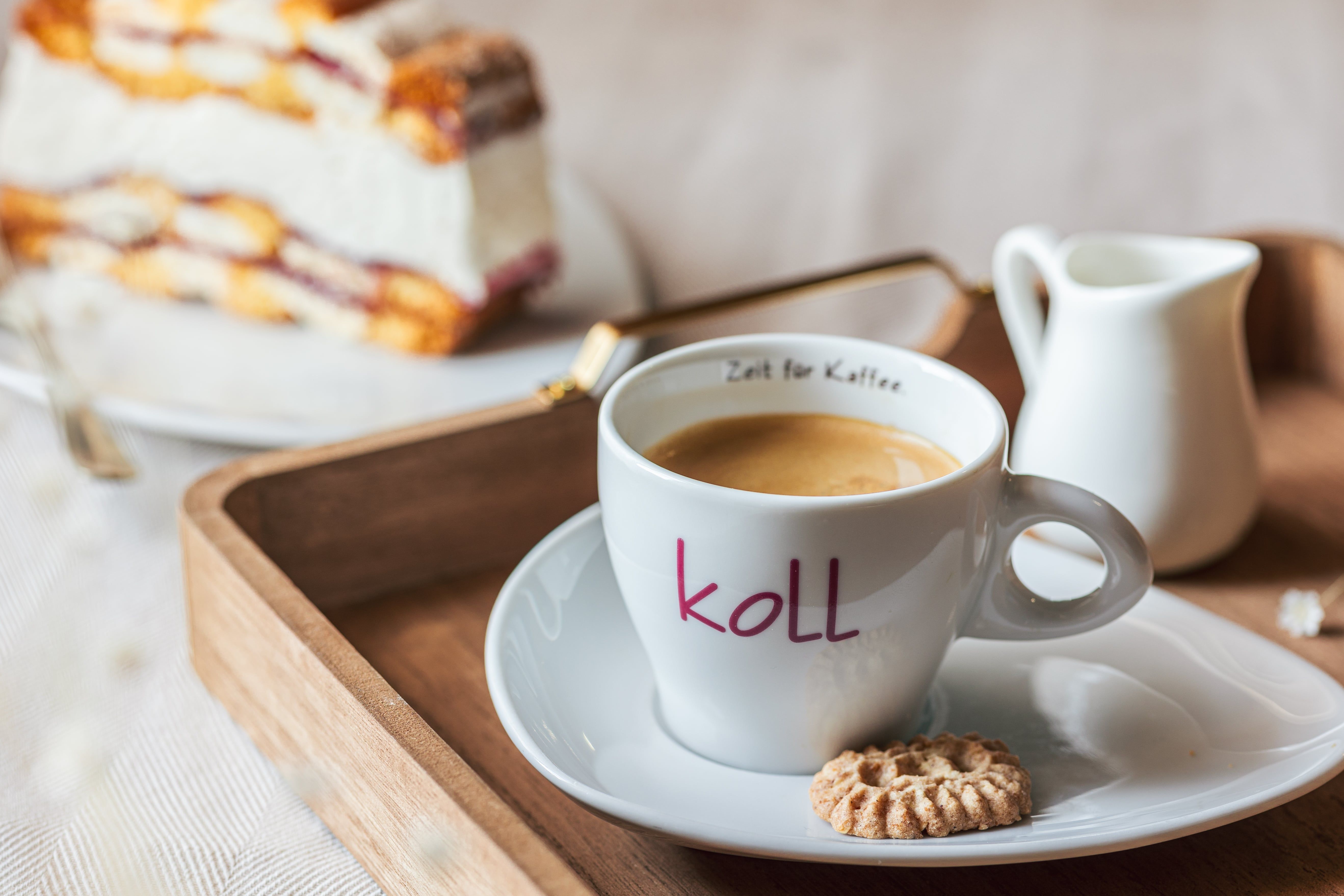 A cup of coffee with a cookie on a tray, a piece of cake in the background.