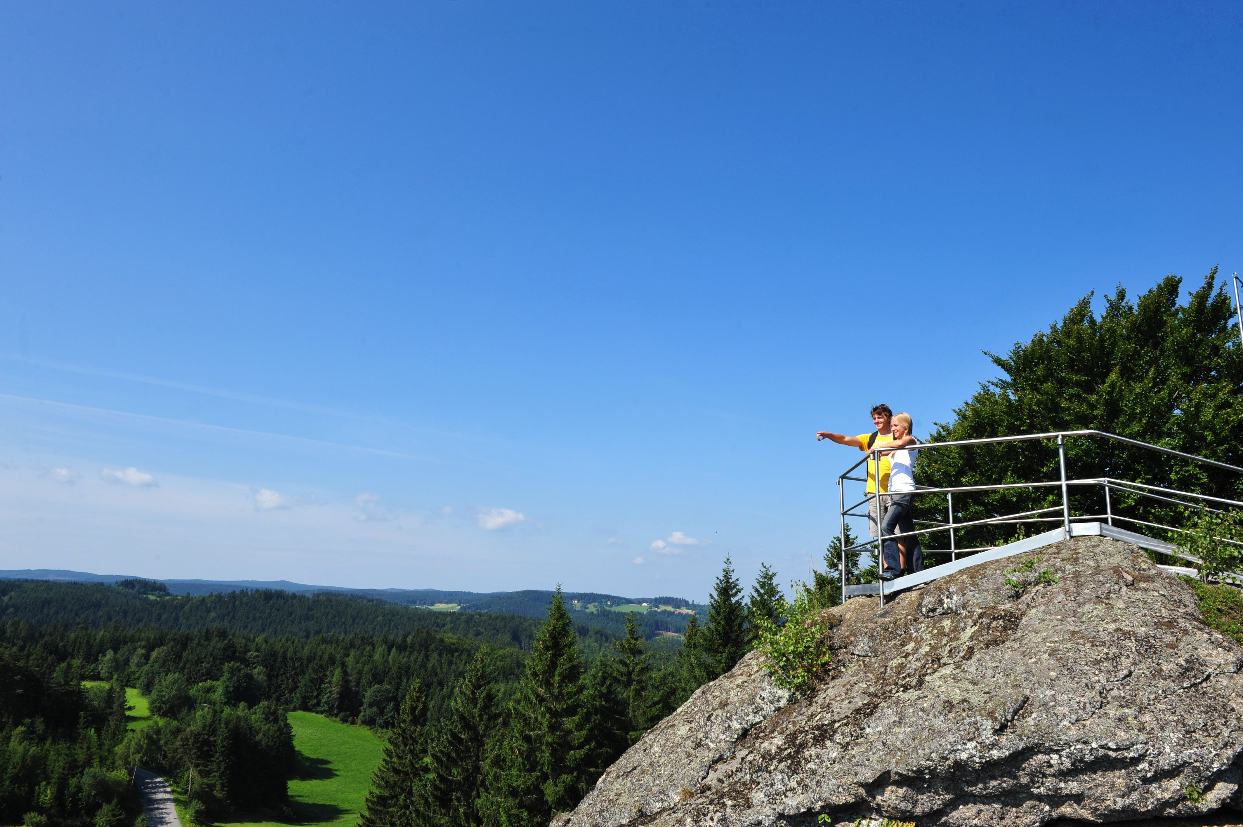 Two people are standing on a lookout point on a rock, gazing into the distance over a wooded landscape.