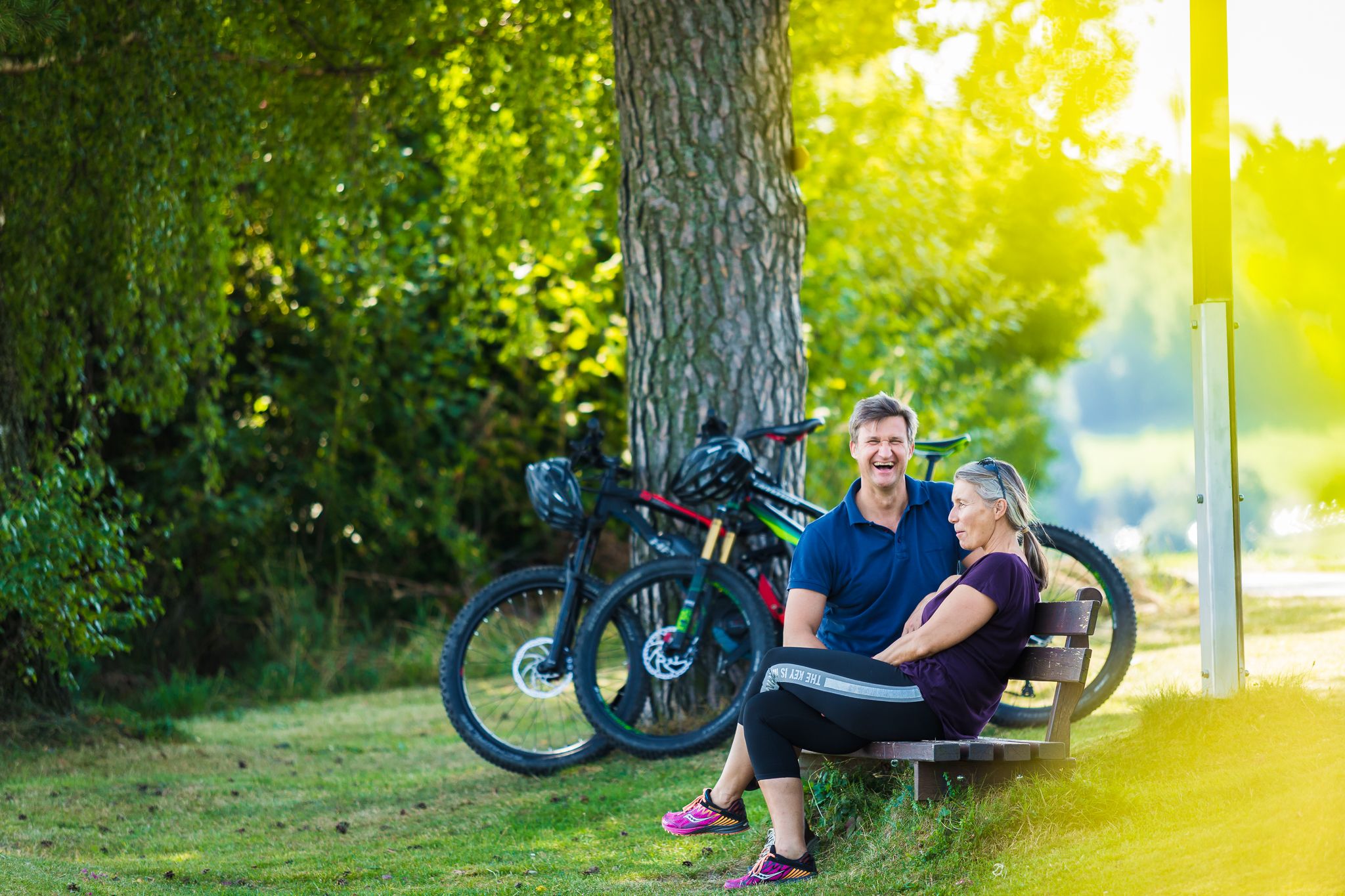 A man and a woman are sitting on a bench in the countryside, two e-bikes are parked next to them.