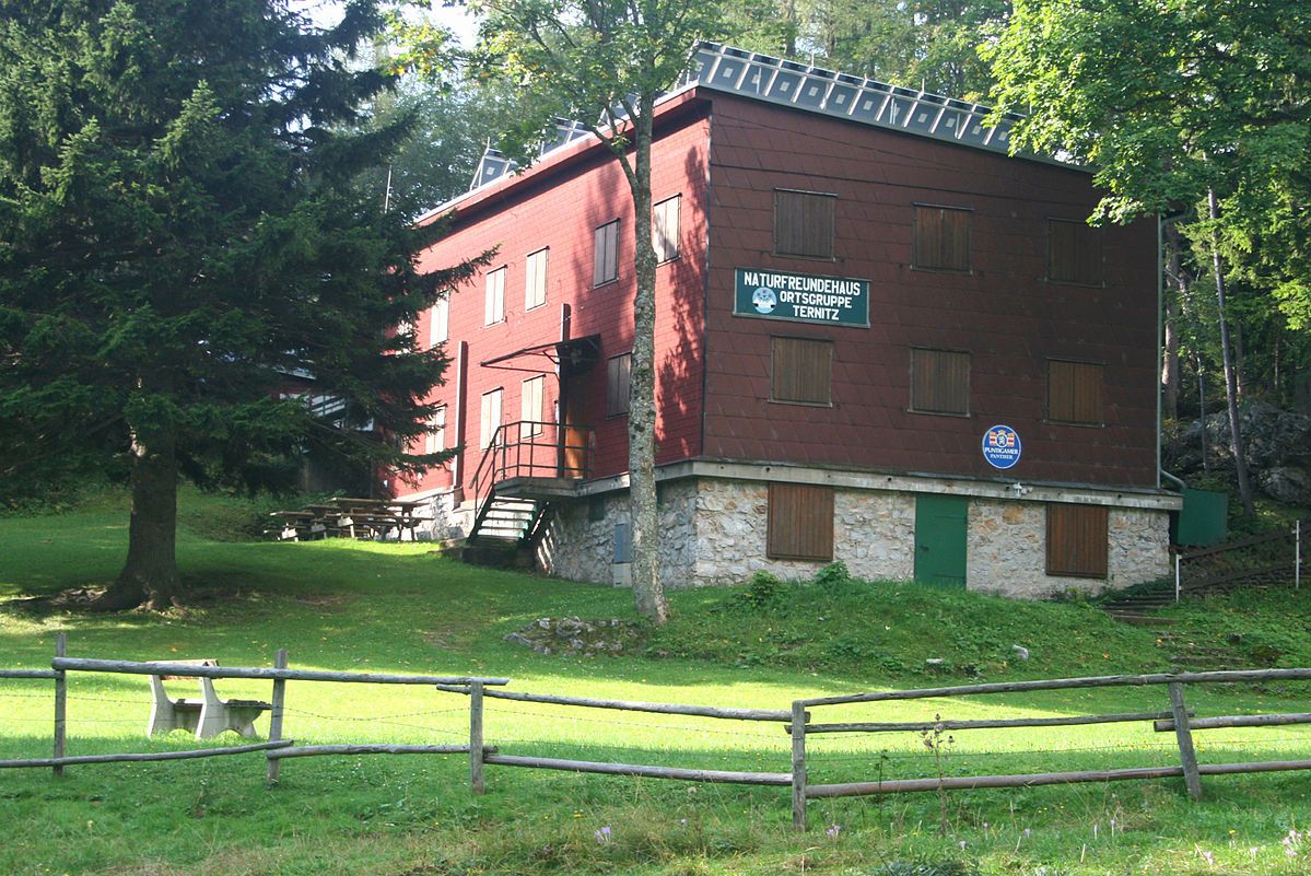 A red building with wooden cladding, surrounded by trees and a meadow, with a sign "Naturfreundehaus Ortsgruppe Ternitz".