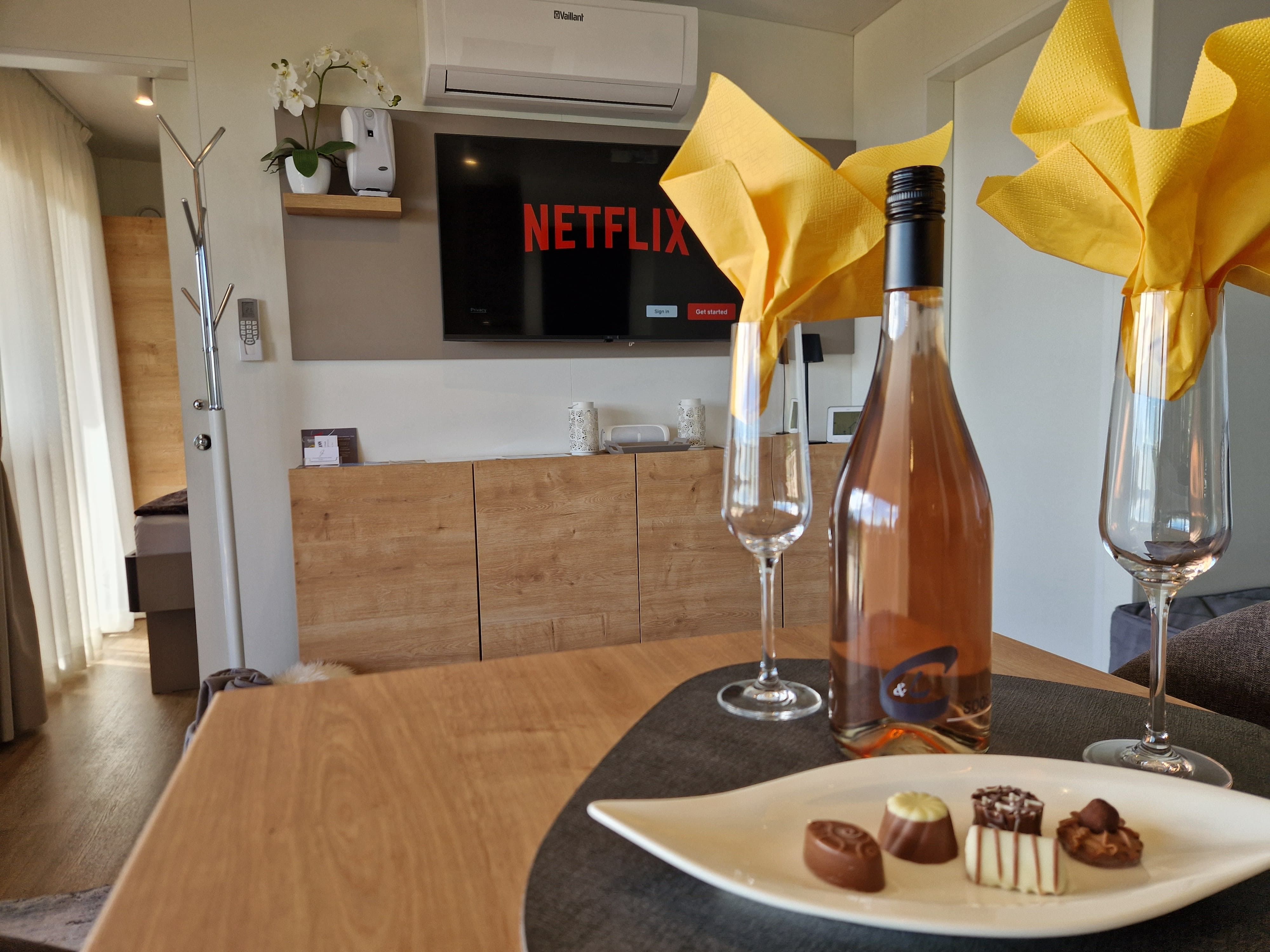 Interior view of a chalet with a bottle of wine, glasses and chocolates on a table, television in the background.
