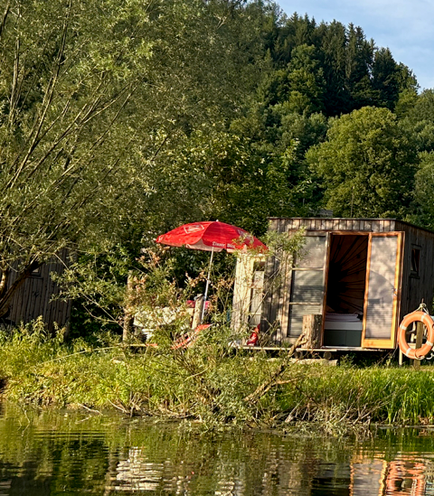 A sleeping pod with a red parasol on the shore of Neubrucker Pond, surrounded by trees.