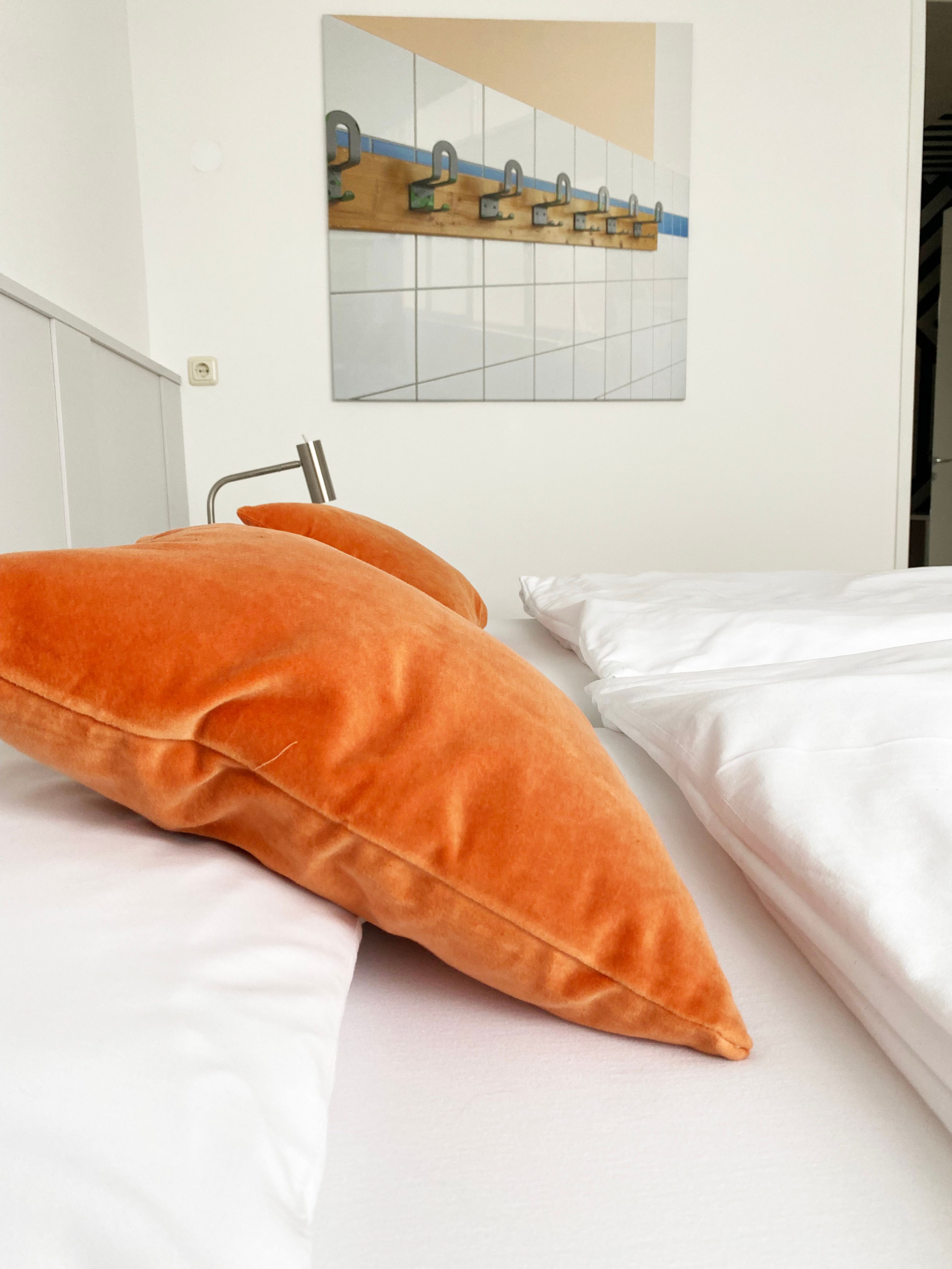 A bed with orange pillows and white bed linen in a guest room. A picture with hooks on tiles hangs on the wall.