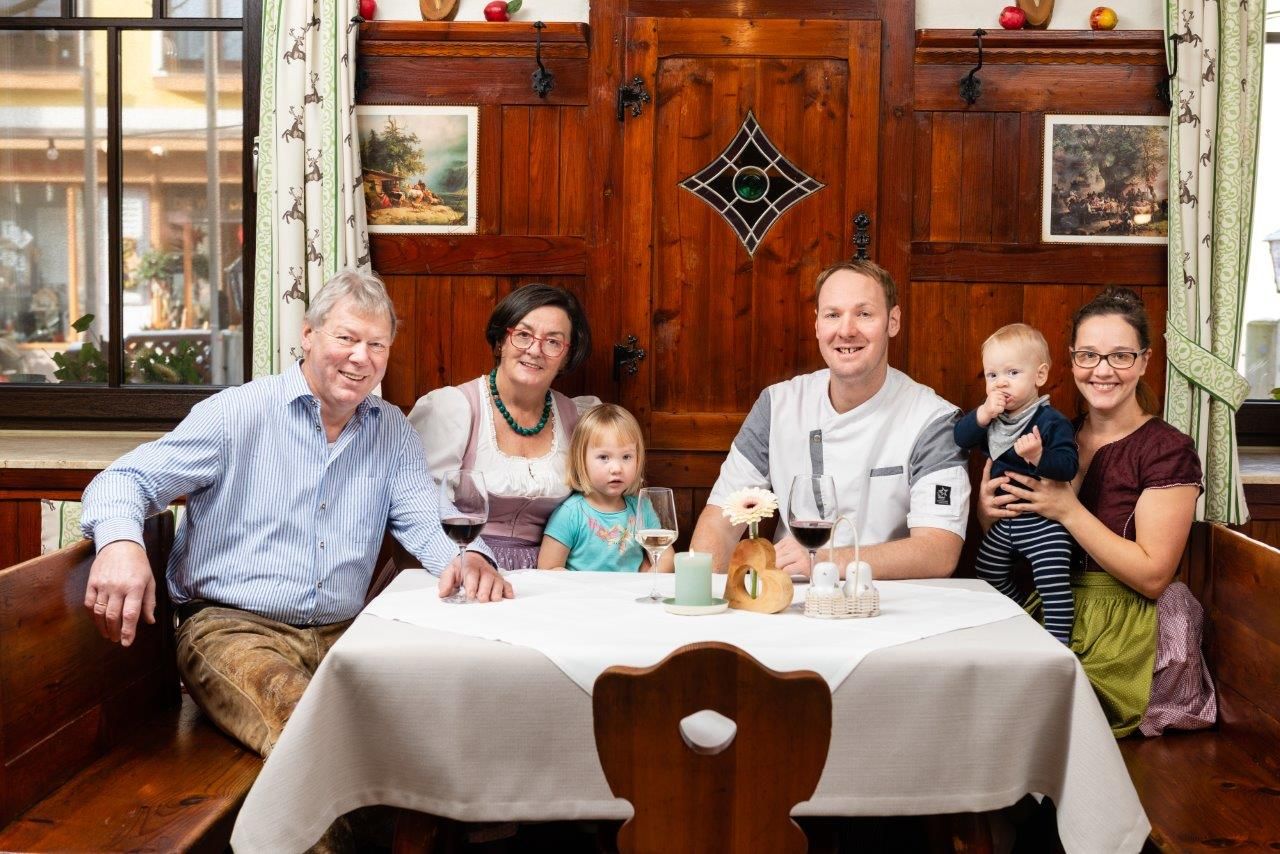 Family sitting at a table in front of a rustic wooden wall. There are 3 wine glasses and decorations on the table.