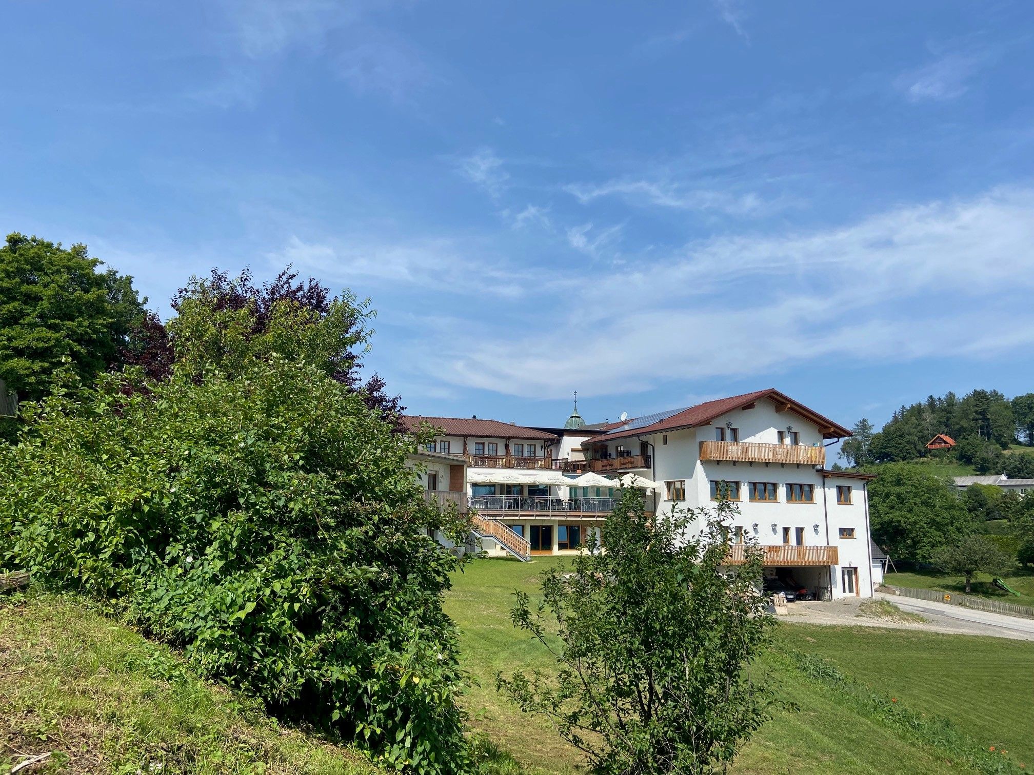 Large building with red roofs and balconies, surrounded by trees and meadows under a blue sky.