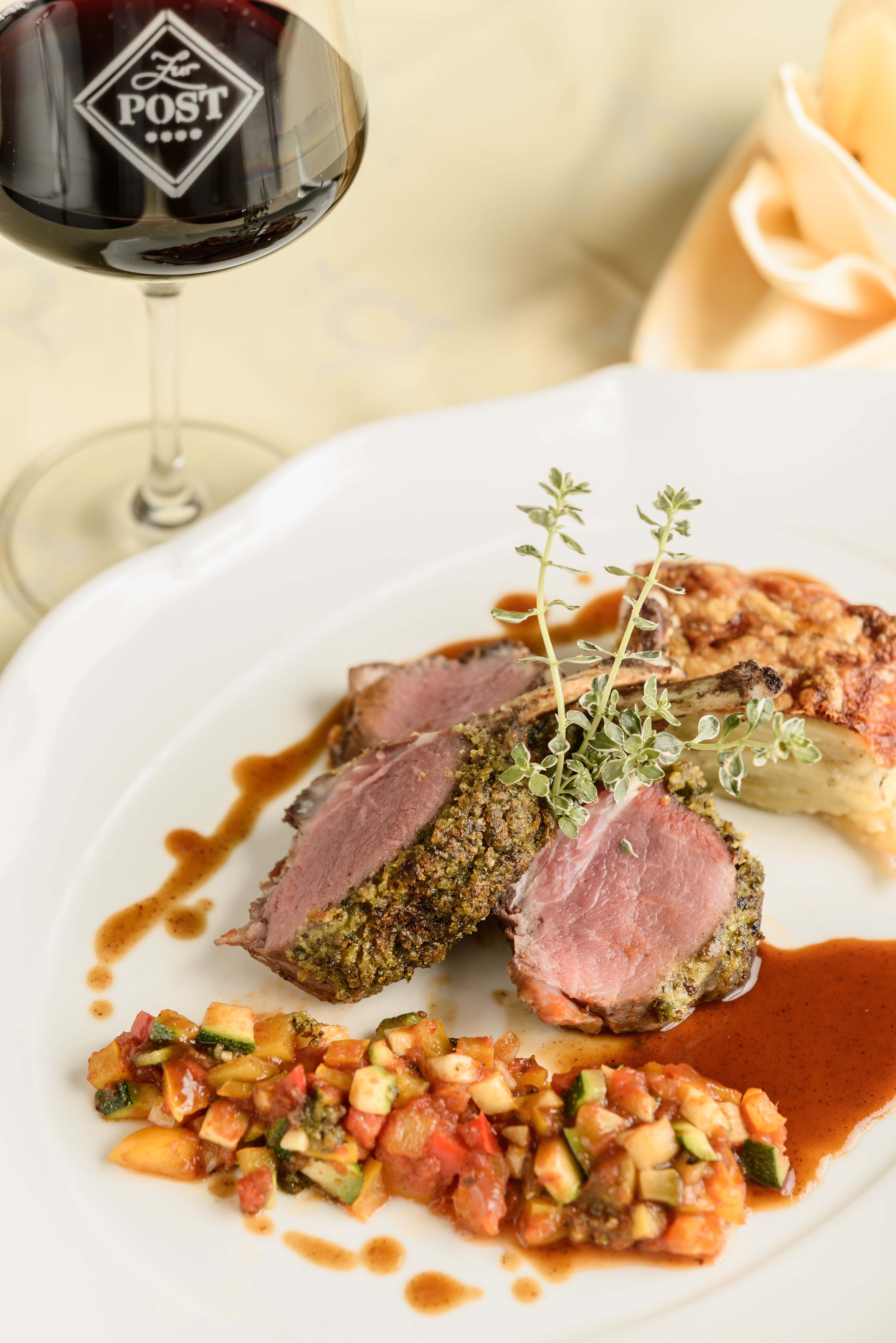 A plate with herb-crusted lamb chops, vegetable garnish and sauce, with a glass of red wine next to it.