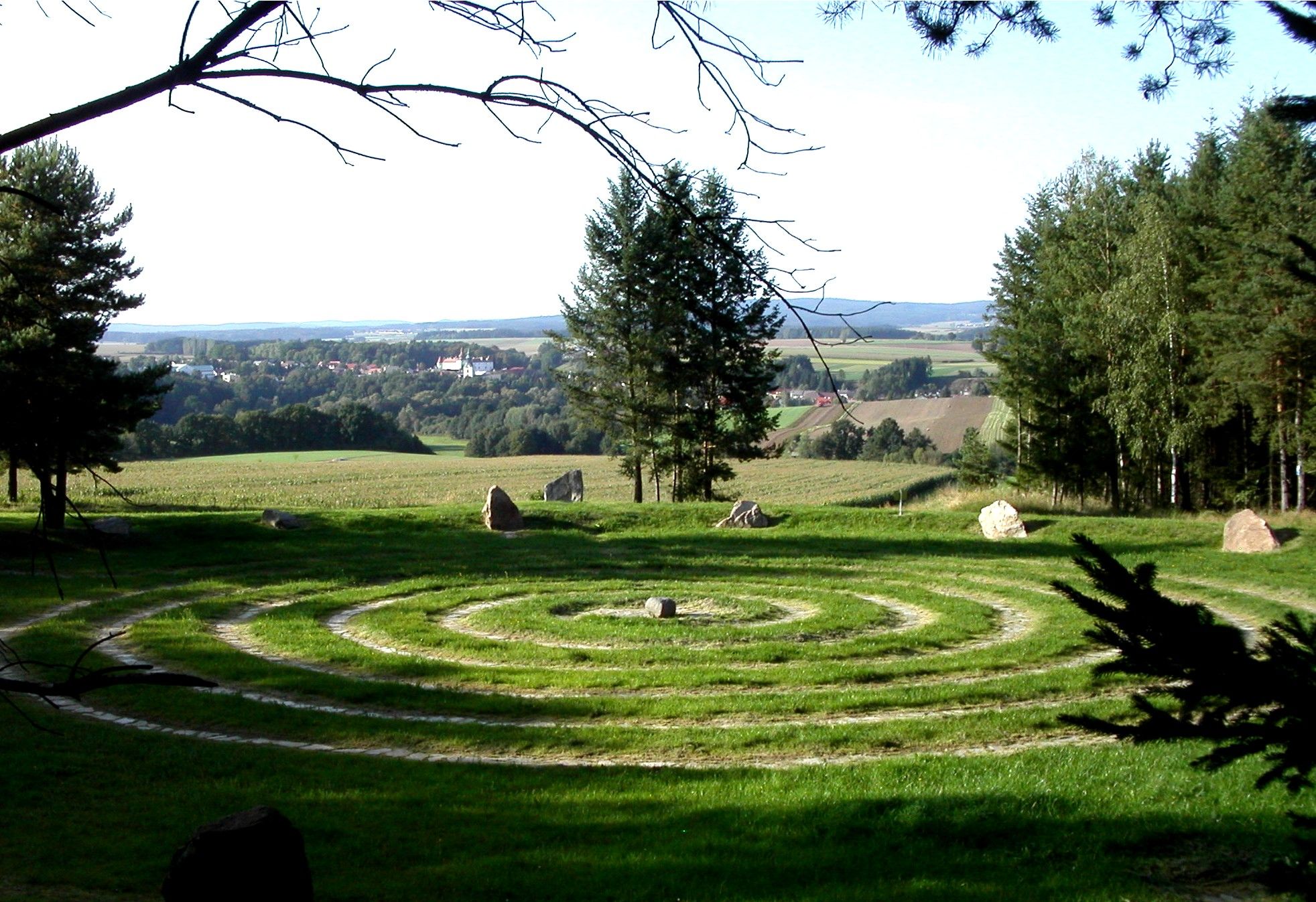 A spiral labyrinth of grass and stones on a meadow with trees and a village in the background.