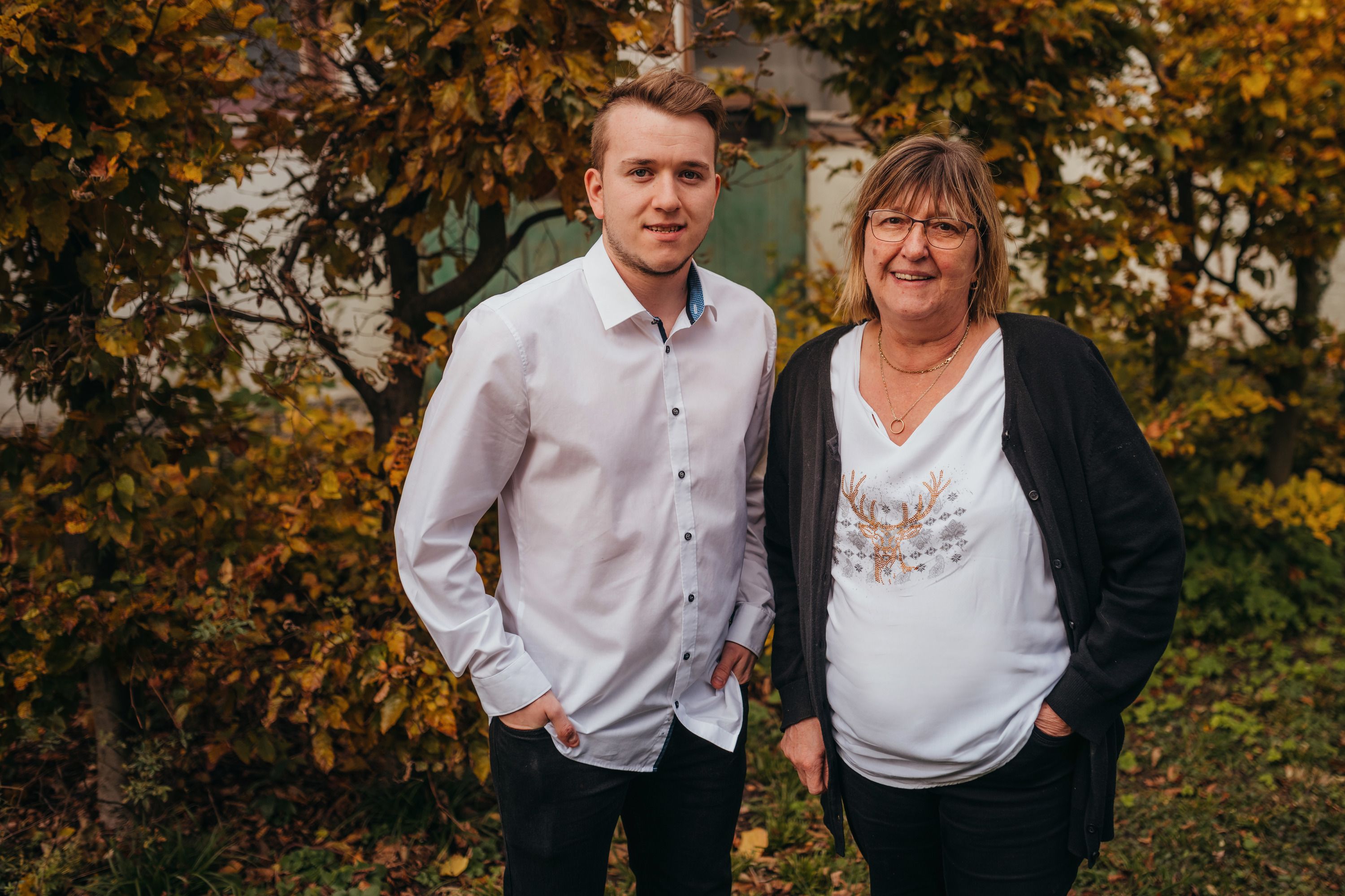 A young man and an older woman stand smiling in front of autumnal trees.