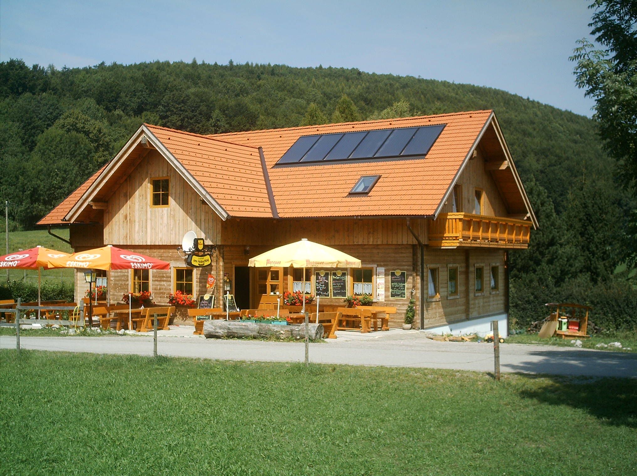 A traditional inn with wooden walls and red roof tiles, surrounded by green countryside and forest.