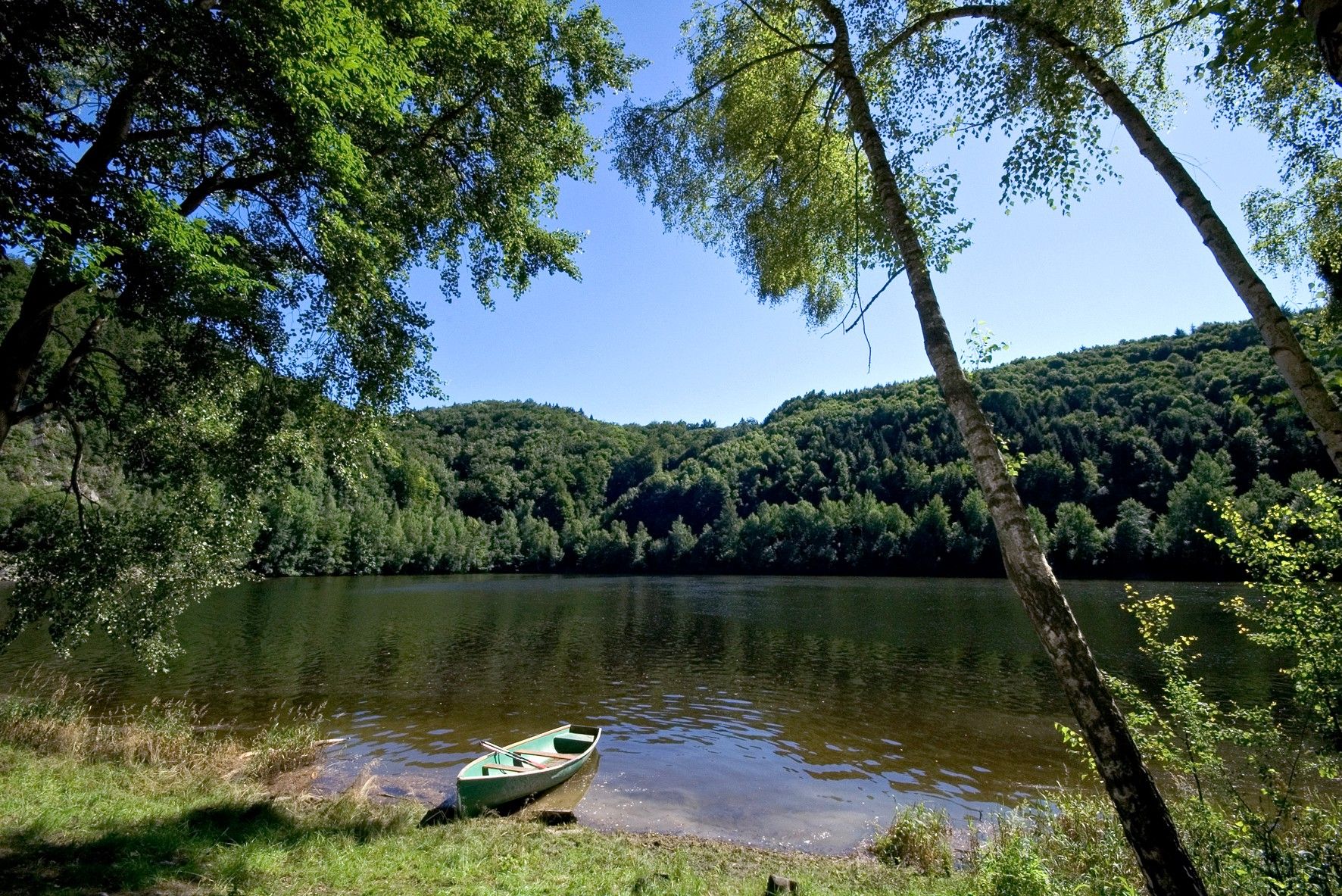 A calm lake with a rowing boat on the shore, surrounded by densely wooded hills and clear skies.