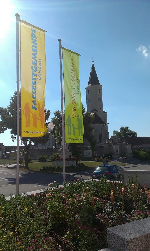 Two colorful flags with the inscription 'Freizeitgemeinde Langau' and 'Do bin i daham' in front of a church.