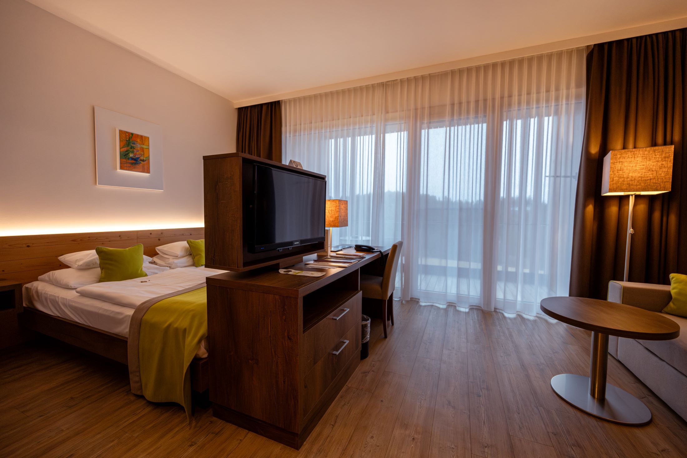 Modern hotel room with double bed, TV, desk and large windows with curtains.