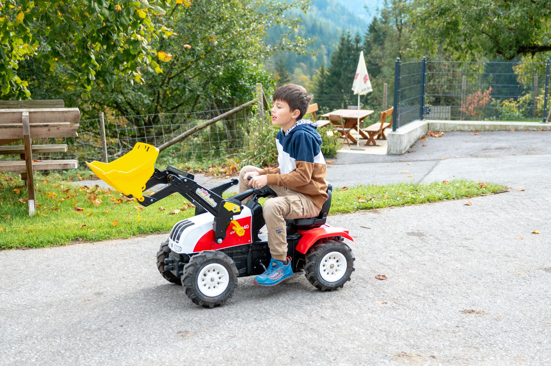 A child is riding a small pedal tractor with a shovel on an outdoor path.