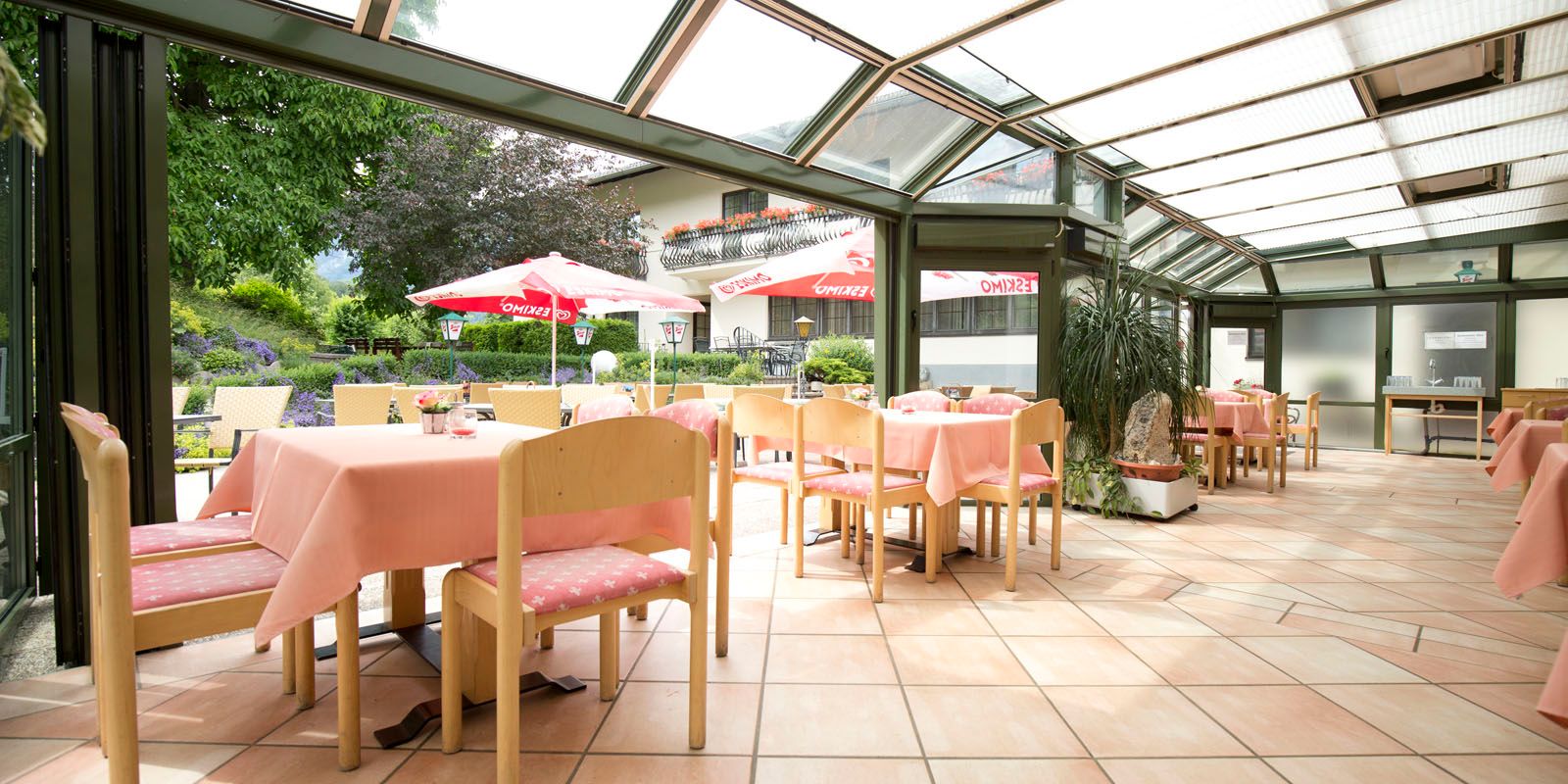 Bright restaurant with glass roof, pink tablecloths and view of the garden with parasols.