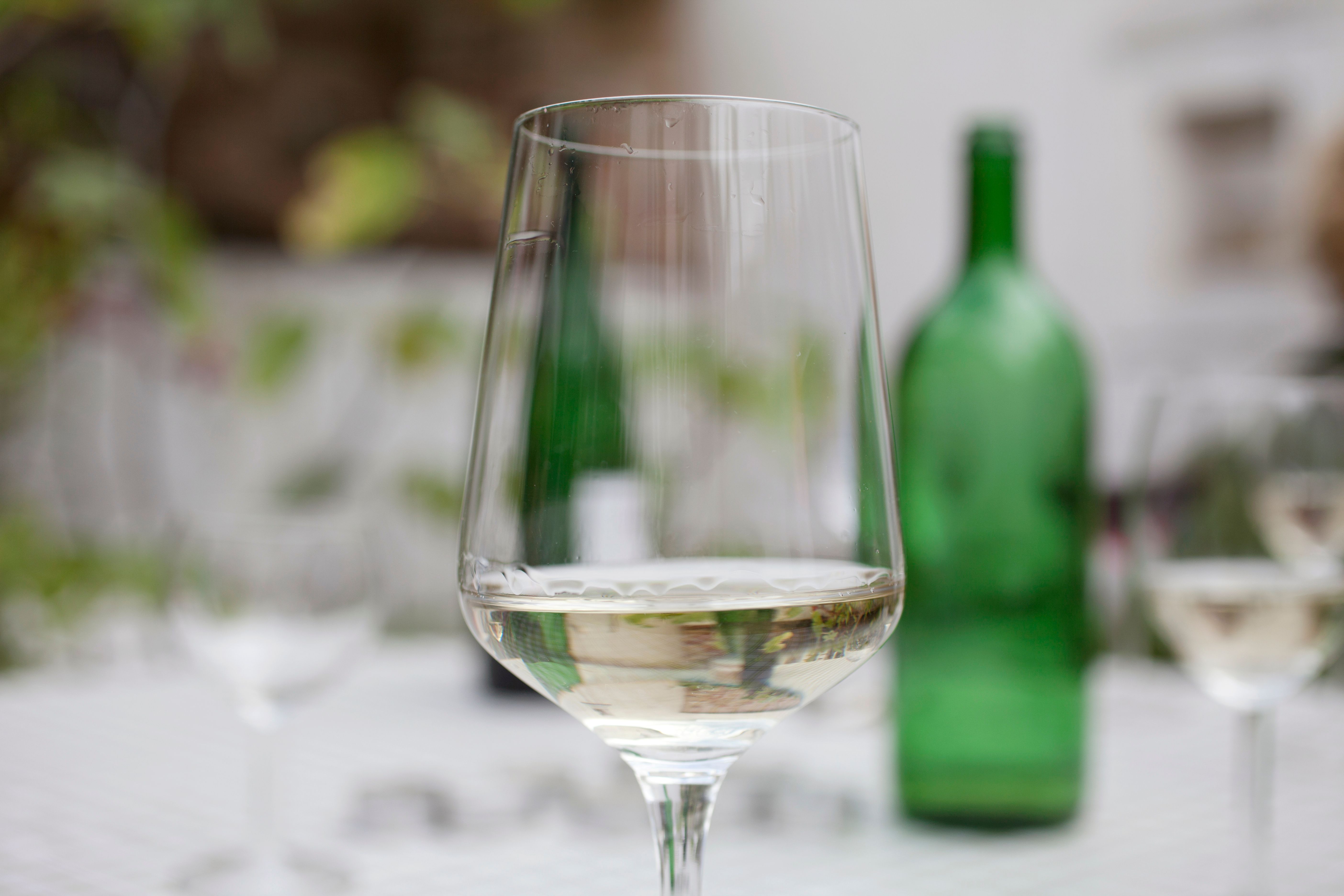 A wine glass with white wine in the foreground, blurred green wine bottle in the background.