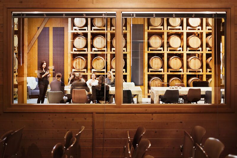 View through a window into a room with whisky barrels and a group of people sitting at a table.