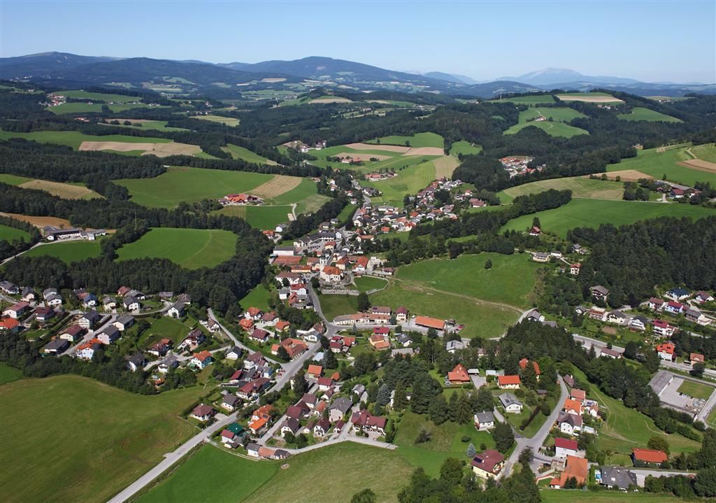 Aerial view of Zöbern, a rural community with houses and fields, surrounded by hills and forests.