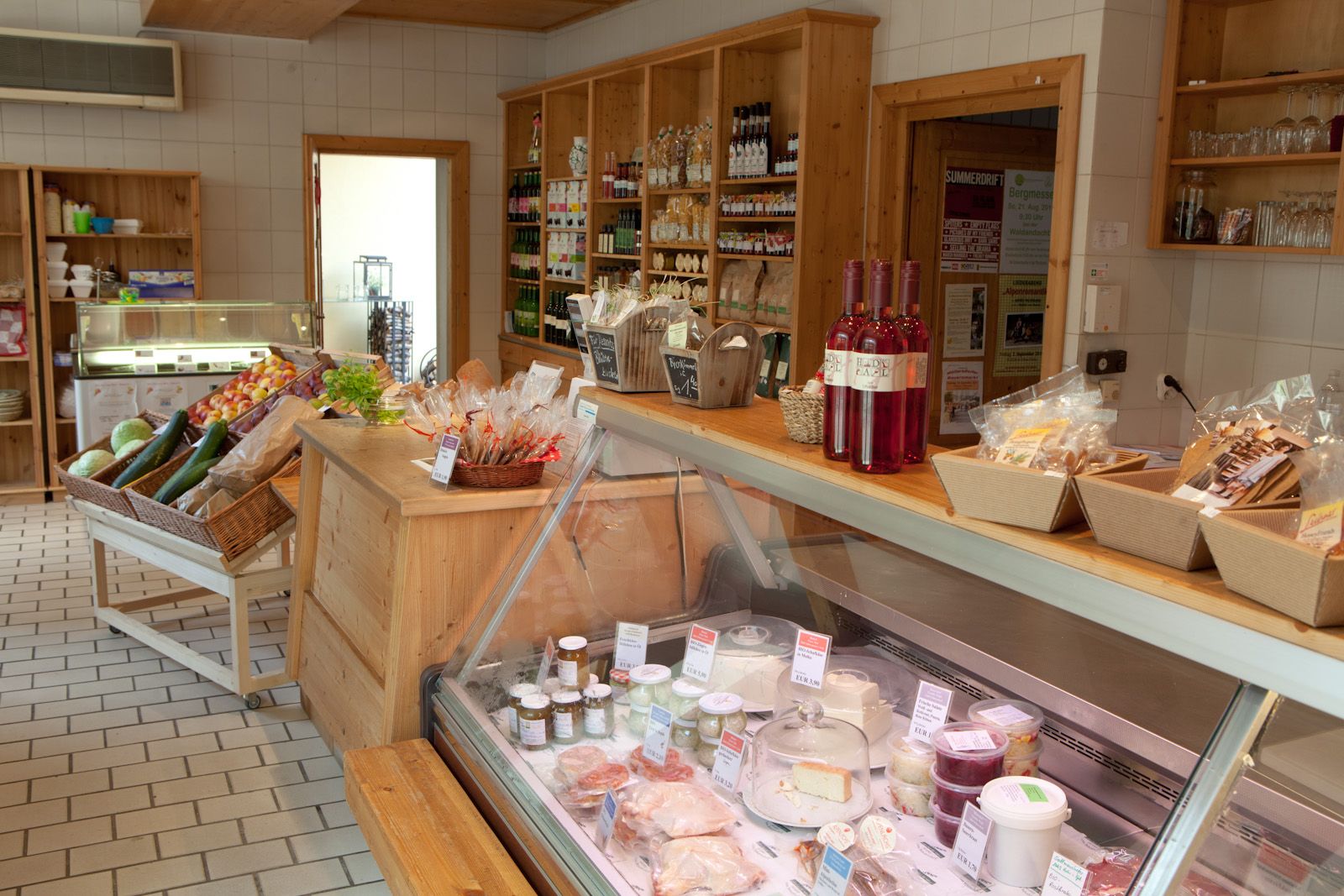 Interior view of a delicatessen with wooden shelves, vegetables, bottles of wine and a refrigerated counter with cheese and spreads.