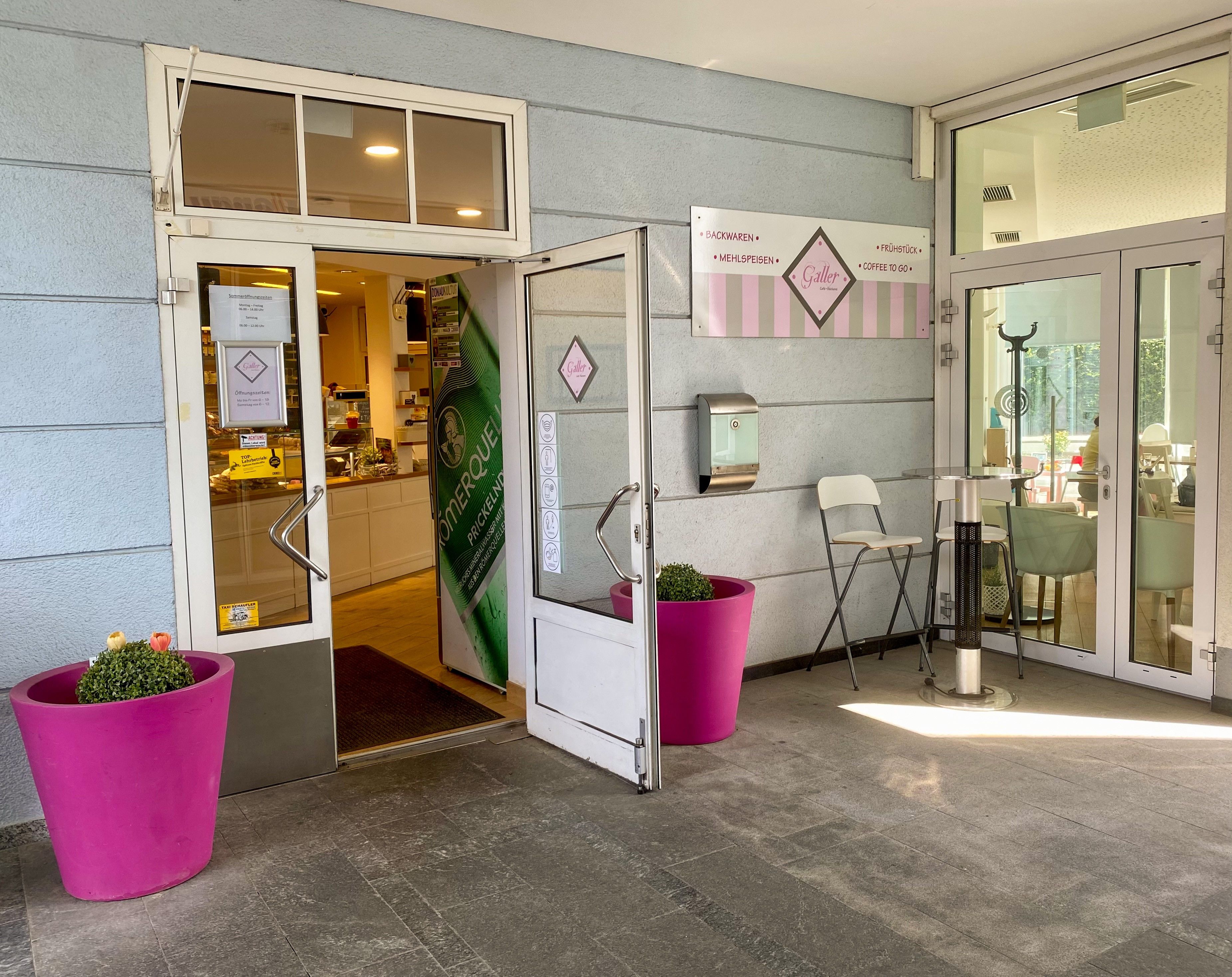 Entrance coffee house klinks and on the right pink flower pots in front of light blue house 