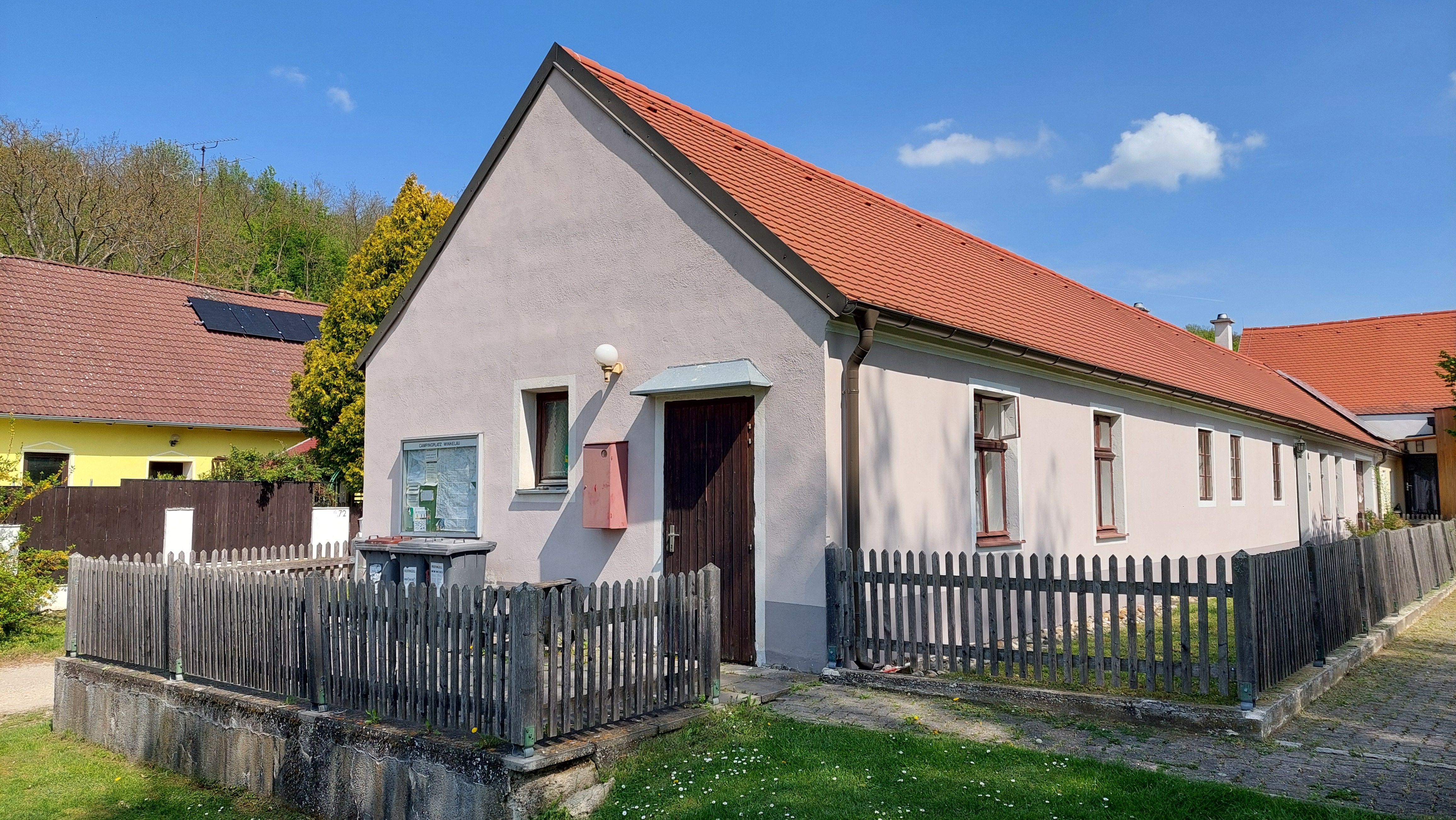 A long, one-story house with a red roof and wooden fence, surrounded by green grass and blue sky.