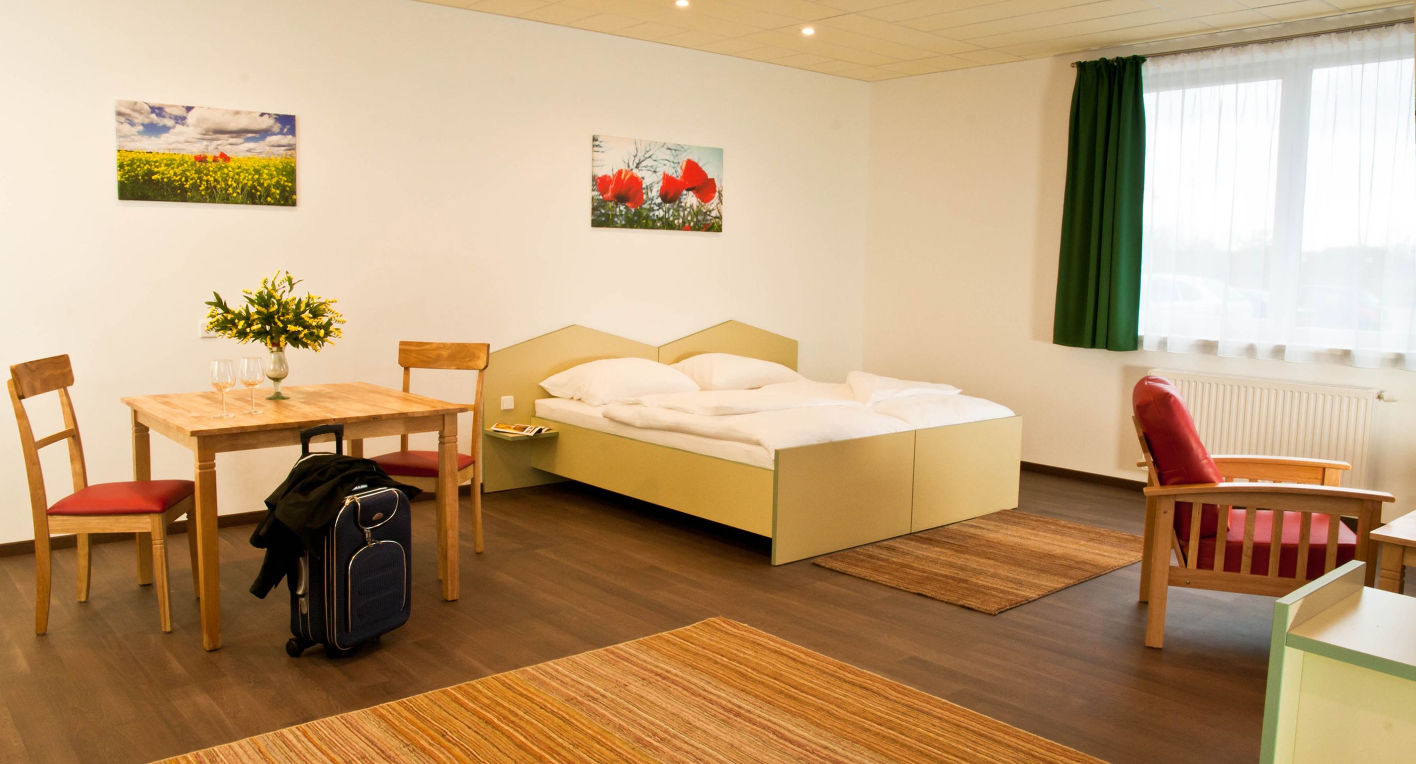 A modern studio with double bed, table and chairs, red armchair and suitcase. Light-colored walls, green curtains and flower pictures on the wall.