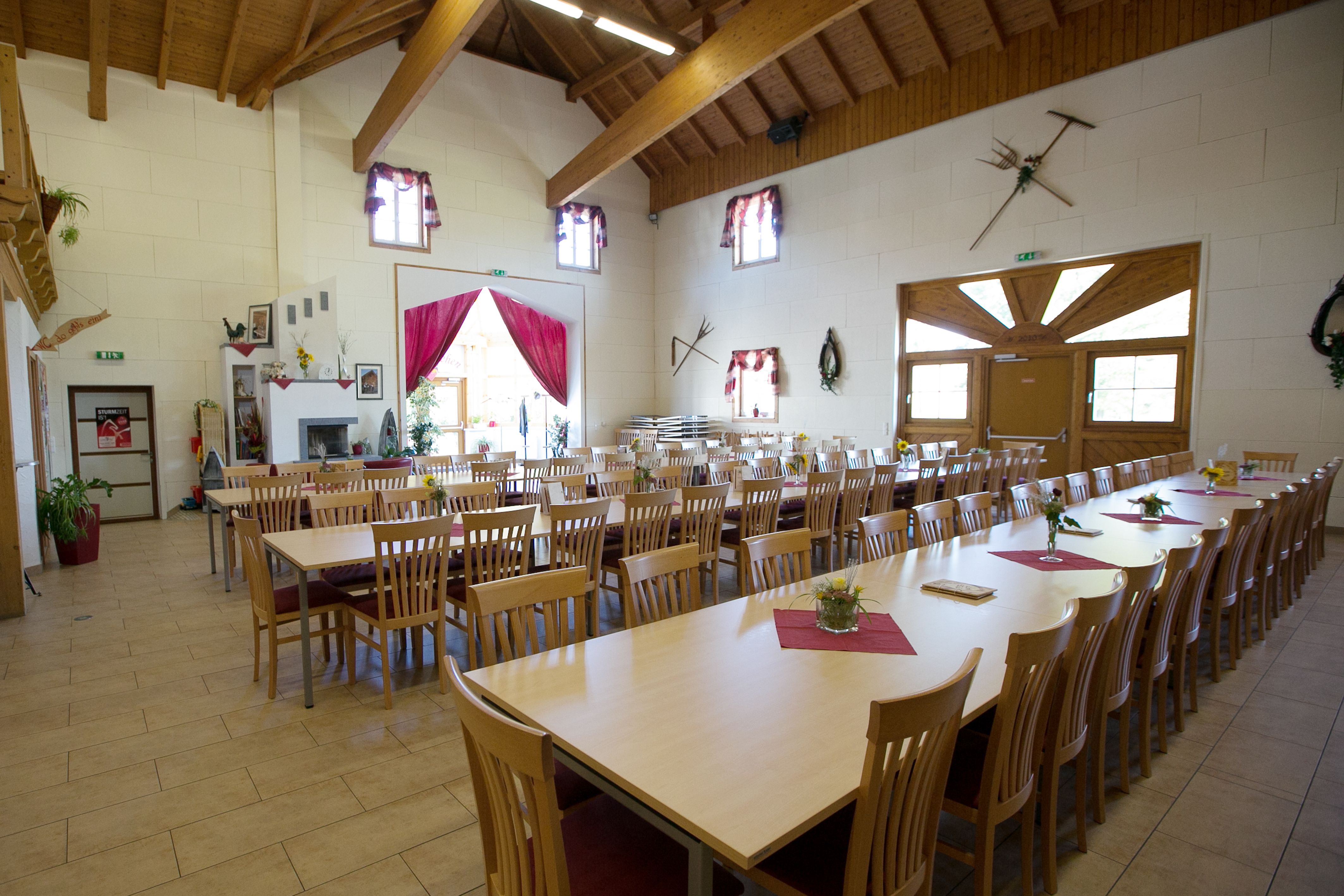 A large, bright room with long wooden tables and chairs, decorated with flowers and red table runners, under a wooden roof.