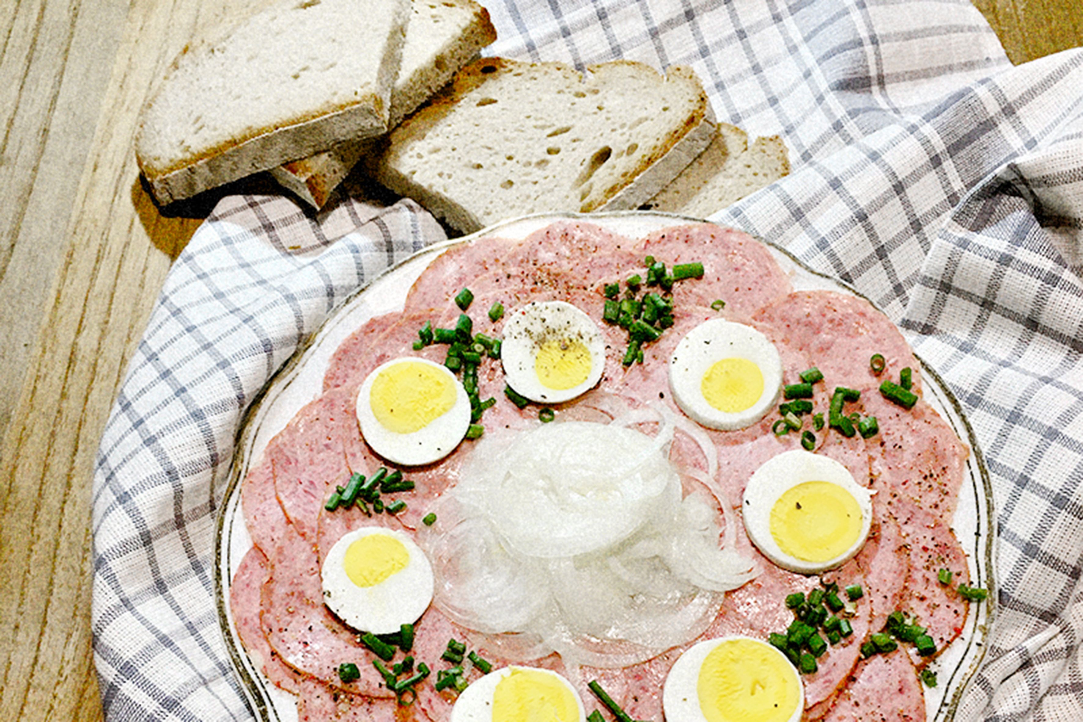 A plate with slices of sausage, hard-boiled eggs, and onions, garnished with chives, next to slices of bread on a checkered cloth.
