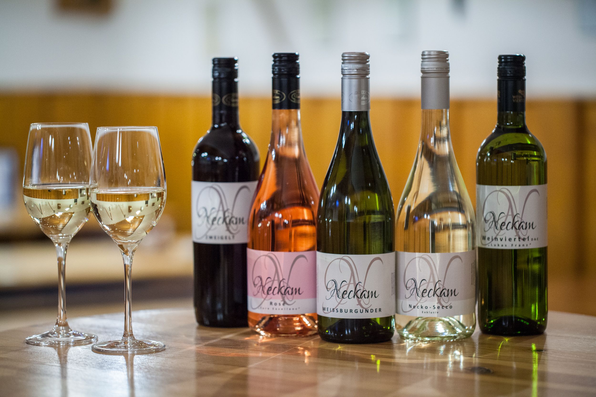 Five bottles of wine and two full wine glasses on a wooden table.