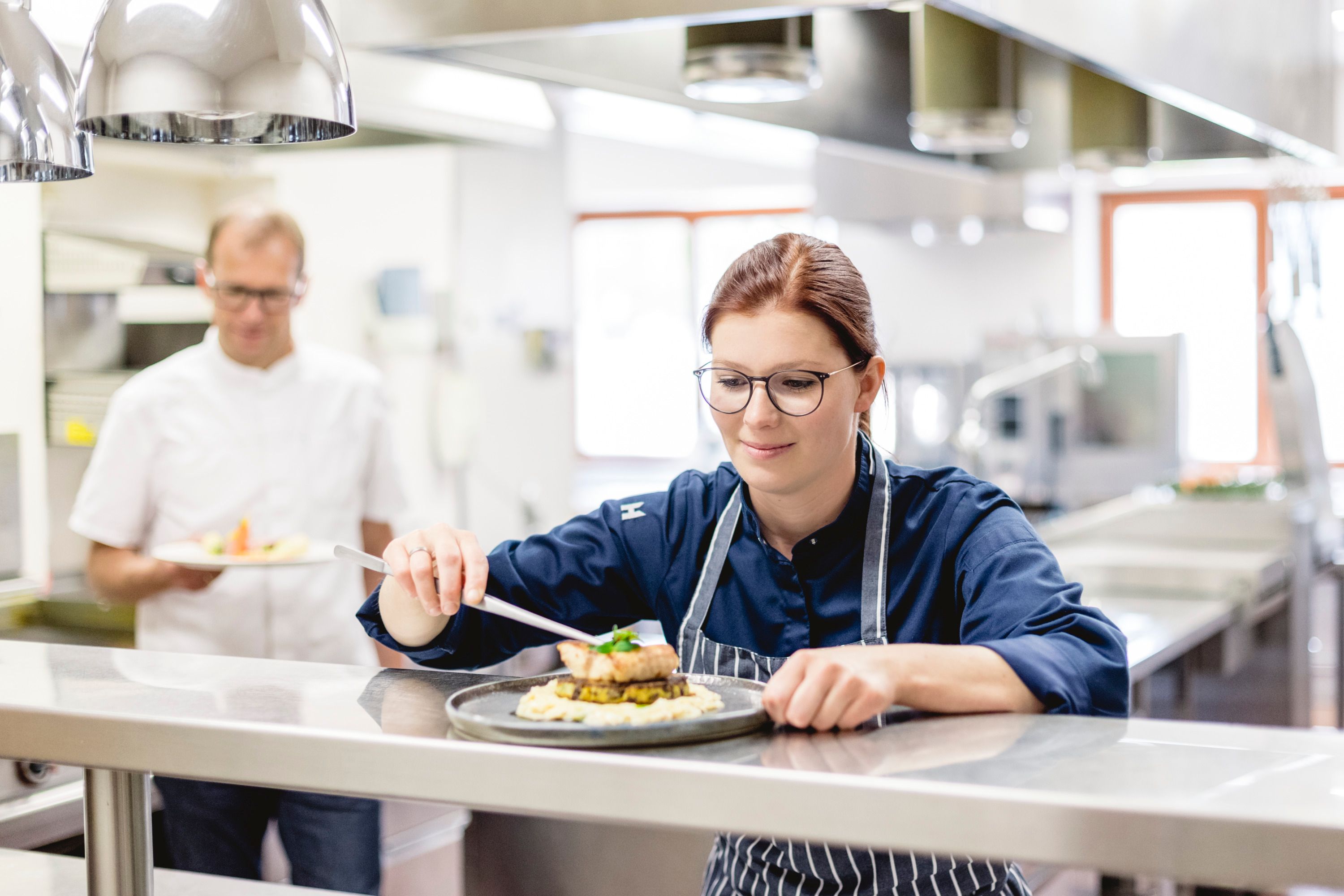 A chef in a restaurant kitchen prepares a dish while a colleague holds another dish in the background.